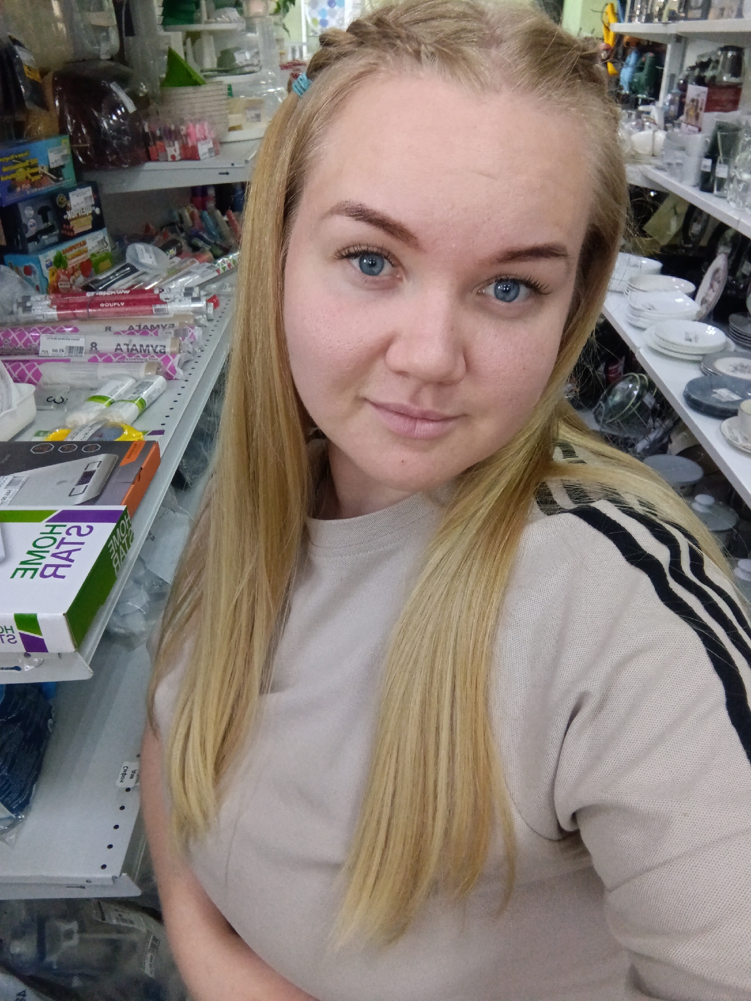 A young woman with fair skin, light blonde hair braided in the front, and blue eyes is taking a selfie. She is wearing a beige sweatshirt with black stripes on the shoulder. She appears to be inside a store, standing between shelves filled with various items. On the left side are boxes and wrapped goods, while the right side displays tableware and glassware. The location appears to be a retail environment within the city of Фоки, Russia.