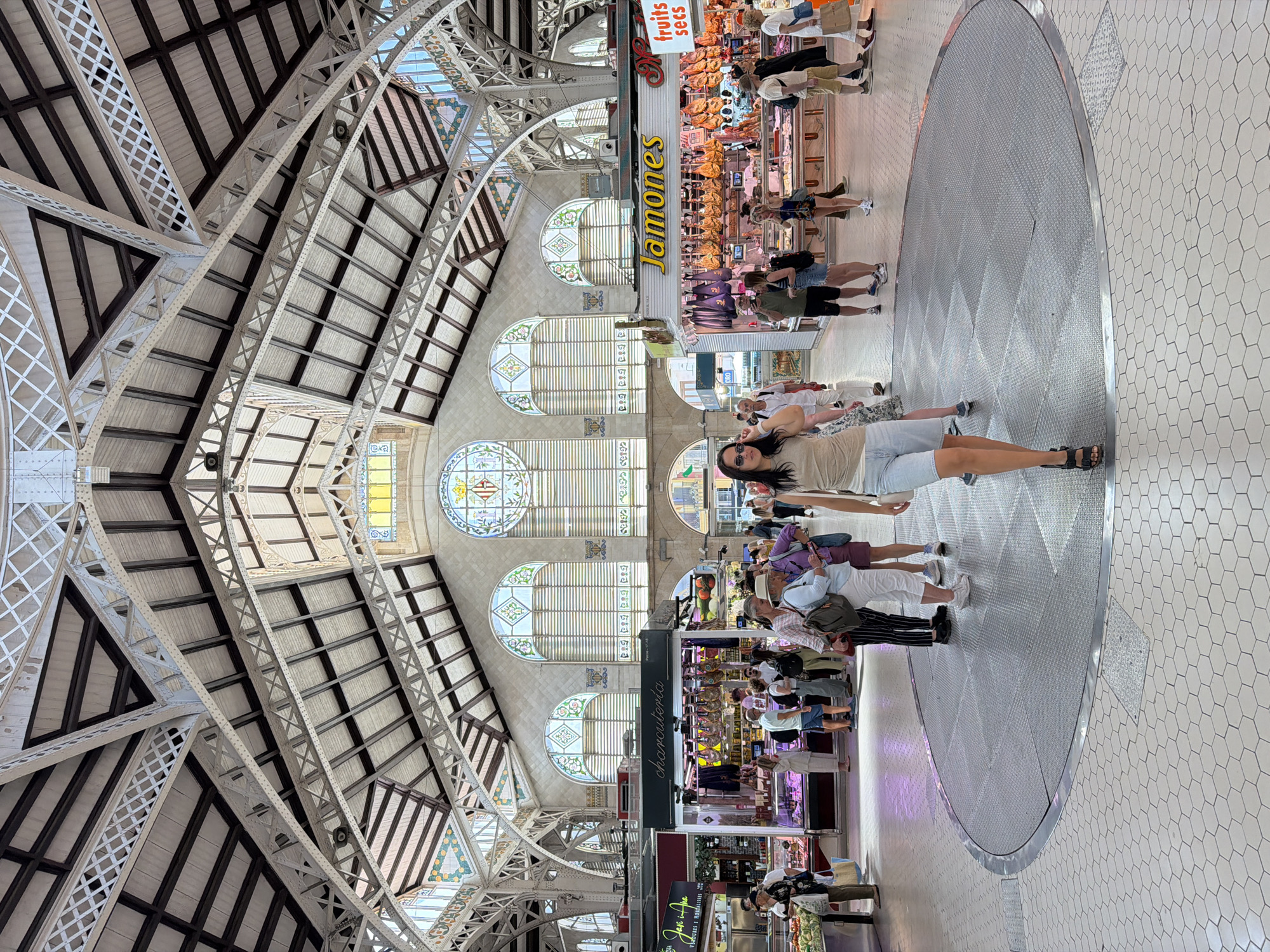 The image shows the interior of a large, well-lit market hall with an ornate wrought-iron ceiling structure. In the foreground, a woman with dark hair wearing a light-colored tank top and denim shorts is walking towards the camera. She is wearing black sandals. She is standing on a circular metal grate, which is surrounded by hexagonal white tiles on the floor. Behind her, other shoppers are visible, some browsing at market stalls displaying meats and produce. The stalls are brightly lit, and the market appears busy. The architecture of the hall features arched windows with stained-glass inserts at the far end. The overall atmosphere is that of a vibrant and historic marketplace.