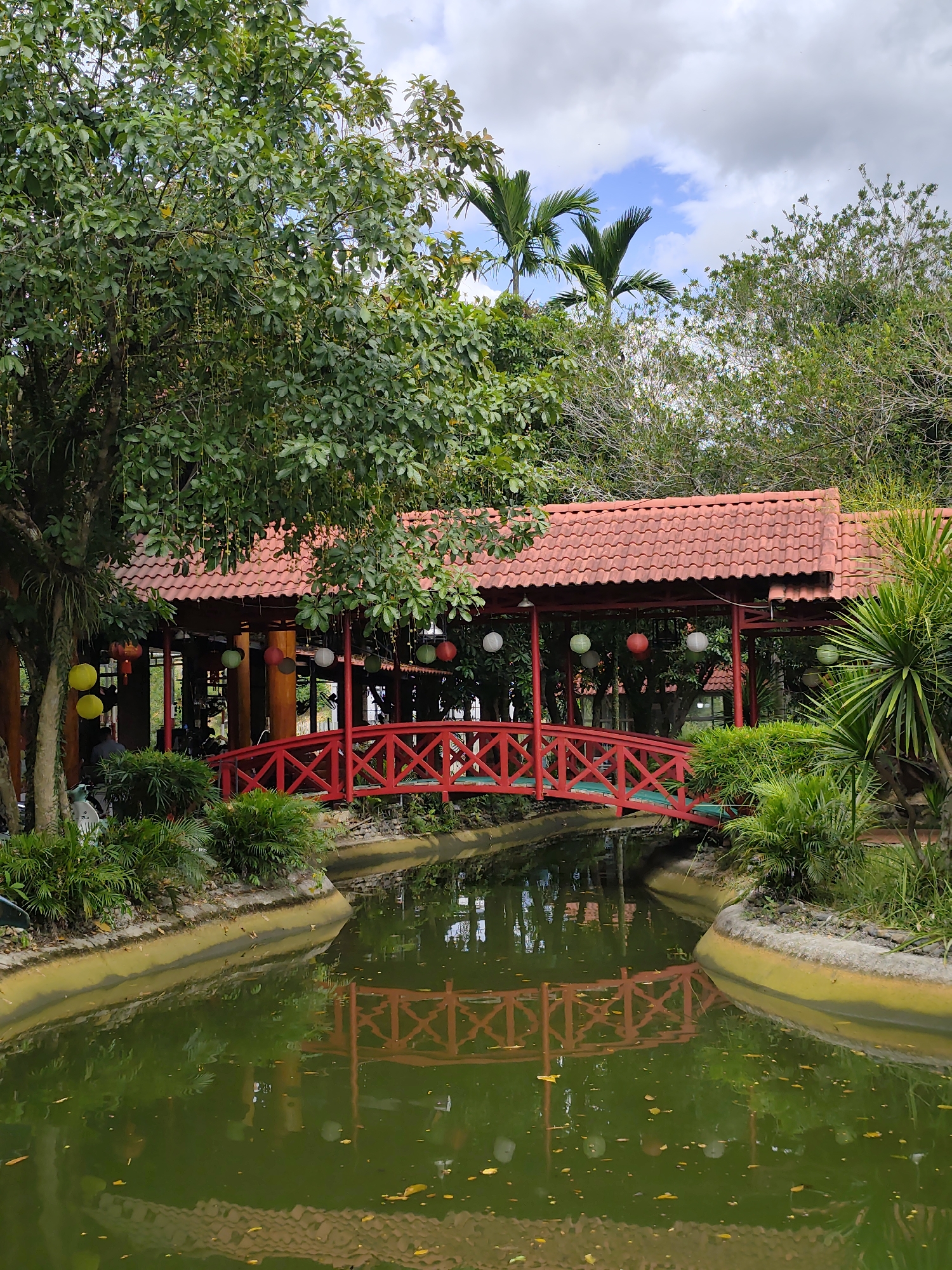 A scenic outdoor view of a red, arched wooden bridge crossing a narrow, tranquil body of water. The bridge, adorned with a decorative railing, leads to a red-roofed building with open sides, suggesting a covered seating area or pavilion. Green foliage, including large trees and smaller bushes, surrounds the waterway and the structure. Several round lanterns, in yellow, white, and red, hang from the building's overhang. The water reflects the bridge and the surrounding greenery, with scattered leaves floating on its surface. The sky above is partly cloudy, with patches of blue visible. The setting appears to be a park or garden, possibly a restaurant or recreational area.