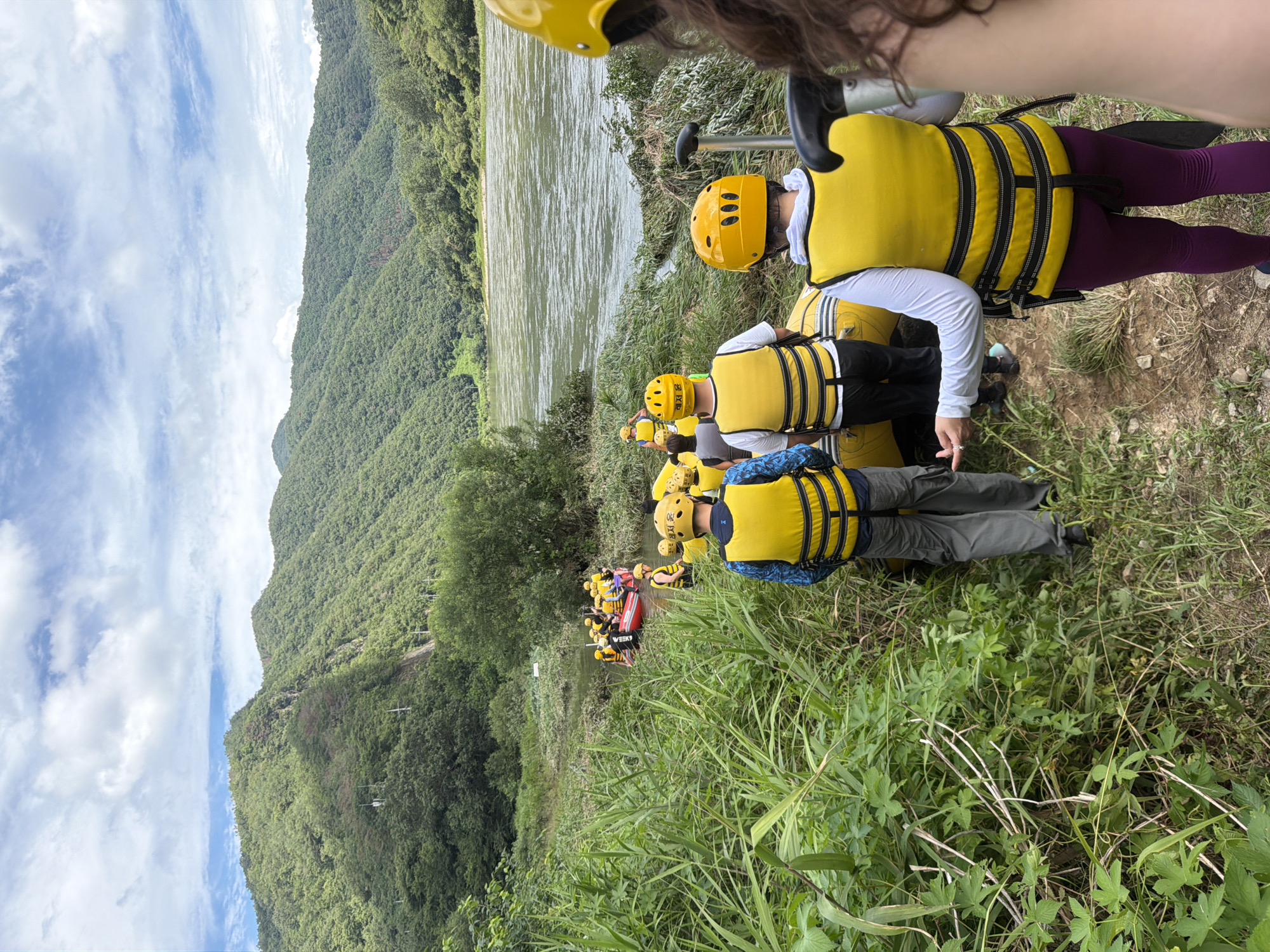 In the Seo-myeon area of South Korea, a group of people are preparing for a rafting excursion. The scene is set by a wide, calm river flowing between lush, green, tree-covered mountains under a sky dotted with white clouds. On the riverbank, amidst tall grasses and foliage, several individuals are visible, predominantly wearing bright yellow life vests and helmets. In the foreground, a person in a yellow vest and grey pants faces away from the camera, gesturing with their left hand. To their right, another person, also in a yellow vest and white long-sleeved shirt, is partially visible. Further into the scene, a line of people in yellow vests and helmets are walking towards and preparing to board inflatable rafts in the shallow water. The rafts are yellow and red, and some are already partially submerged. The environment is natural and undeveloped, with the river and mountains forming a serene backdrop for the recreational activity.