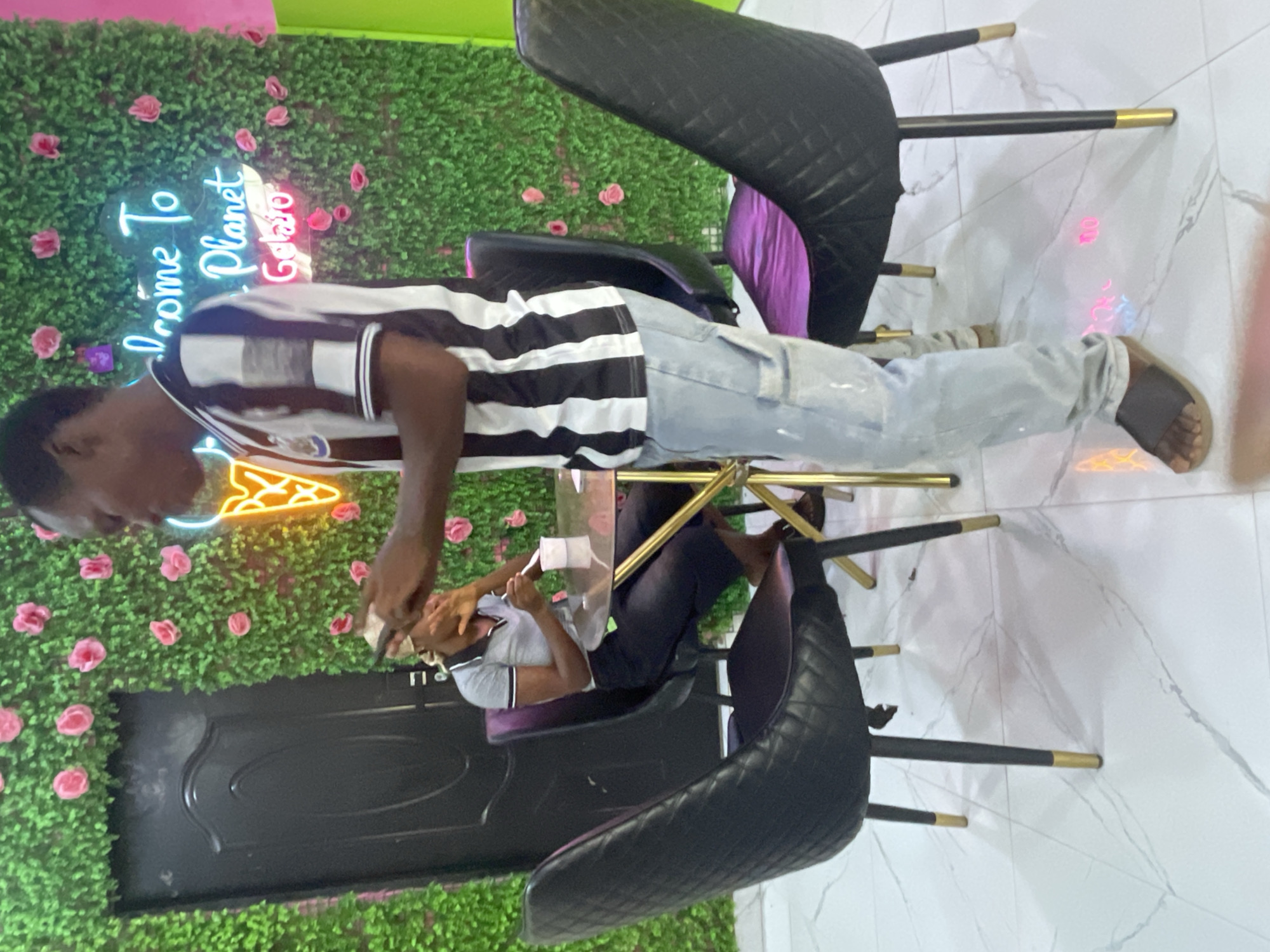 In Jalingo, Nigeria, a man wearing a black and white striped football jersey and light blue jeans stands near a table, looking at his phone. He is wearing dark brown slides. Seated at the table, a woman in a grey polo shirt and dark pants is eating from a white container with a spoon. The scene is set against a wall covered in artificial green foliage and pink artificial flowers. A neon sign with the words "Welcome To Planet Gelato" is visible. The flooring is white marble tile with grey veining. Black chairs with purple cushions are positioned around the table.