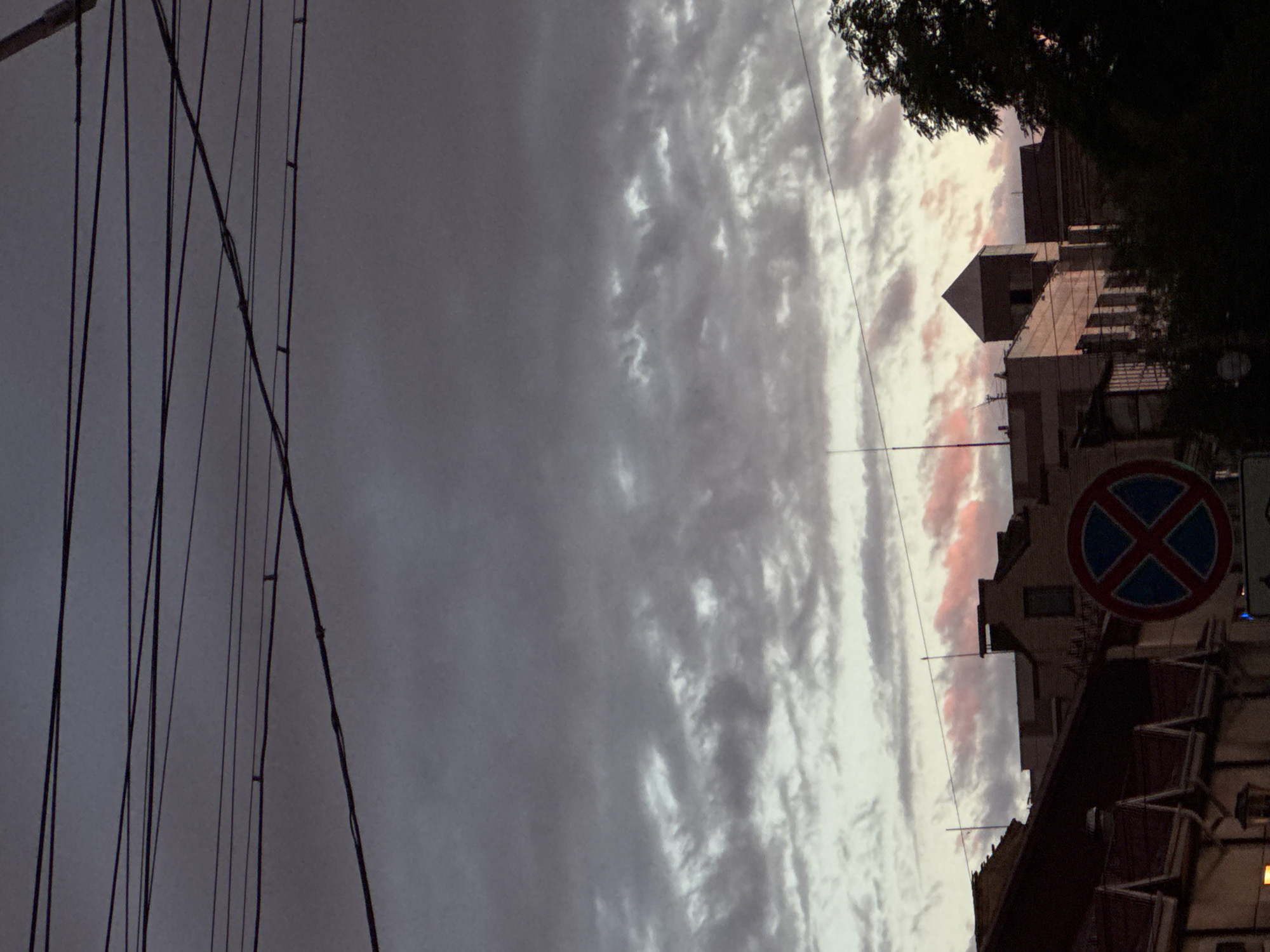 A low-angle, medium shot captures a cityscape with a dramatic sky at dusk. Numerous power lines crisscross the frame against a backdrop of dark, cloudy skies that are tinged with pale pink and white hues near the horizon. The tops of buildings are visible below the sky, with architectural details including peaked roofs and a square tower. A circular traffic sign with a blue background and a red cross is prominently displayed in the lower center of the image. The overall scene is dark and moody, suggesting an evening setting in an urban environment.