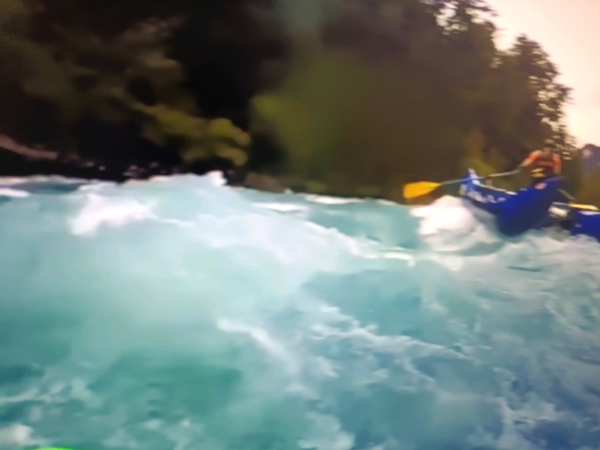 The image captures a scene of whitewater rafting on a turbulent river. In the foreground, the bright blue water churns with white foam, indicating rapids. A blue raft is visible, carrying at least one person wearing an orange life jacket. This individual is using a yellow paddle to navigate the water. The background features a dense, dark green forest bordering the river, with some lighter areas suggesting sunlit foliage or open sky. The overall impression is one of adventure and active recreation in a natural, wilderness setting. There is no discernible connection to the city of Netanya, Israel, in this image.