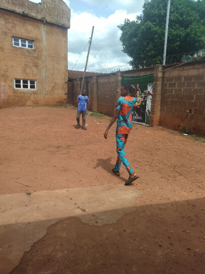 The image shows two young men in an open-air courtyard in Anyigba, Nigeria, located at latitude: 7.503, longitude: 7.175. One man, wearing a blue t-shirt and blue jeans, is walking away from the camera. The other man, wearing a multi-colored shirt and shorts, is walking towards the camera. Both men are walking on a dirt path. There is a brown brick building to the left of the frame and a brick wall to the right of the frame. The wall is covered with a green poster. There are several trees in the background. The ground is covered in dirt. The scene is bright, with no shadows. The sky is clear and blue, with some white clouds. The atmosphere is calm and peaceful.
