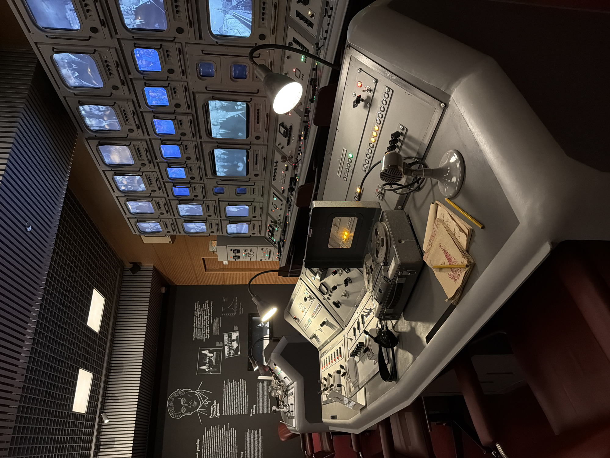 This image captures a dimly lit, retro-futuristic control room or media studio, likely a museum exhibit in Moscow, Russia. The central focus is a large, gray console stretching across the foreground, equipped with numerous buttons, switches, dials, and indicator lights, suggesting a complex operational interface. Several adjustable task lamps illuminate parts of the console, with one shining brightly. A vintage microphone and a reel-to-reel tape recorder are prominent on the desk, alongside some folded papers or notebooks and a pencil. Red upholstered chairs line the console, indicating operator positions.
Above the console, an impressive array of small, square, monochrome television monitors is embedded into the ceiling or upper wall, displaying various black-and-white or blue-tinted images, including cityscapes and historical footage of people. The overall aesthetic, including the CRT screens and analog controls, points to technology from the mid-20th century.
The left wall features a dark panel with white Cyrillic text and illustrations, functioning as an informational display. Notable text visible on this panel includes "ВЫСОЦКИЙ ВЛАДИМИР СЕМЁНОВИЧ" (VYSOTSKY VLADIMIR SEMYONOVICH), linking the exhibit to the famous Soviet cultural figure. Adjacent to this display, the wall is covered with a patterned, possibly acoustic, panel with two recessed rectangular lights.
No people are present, but the setup implies activities such as monitoring, broadcasting, or managing a complex system. The controlled lighting and detailed historical equipment create an immersive experience, showcasing a significant era of technological development.