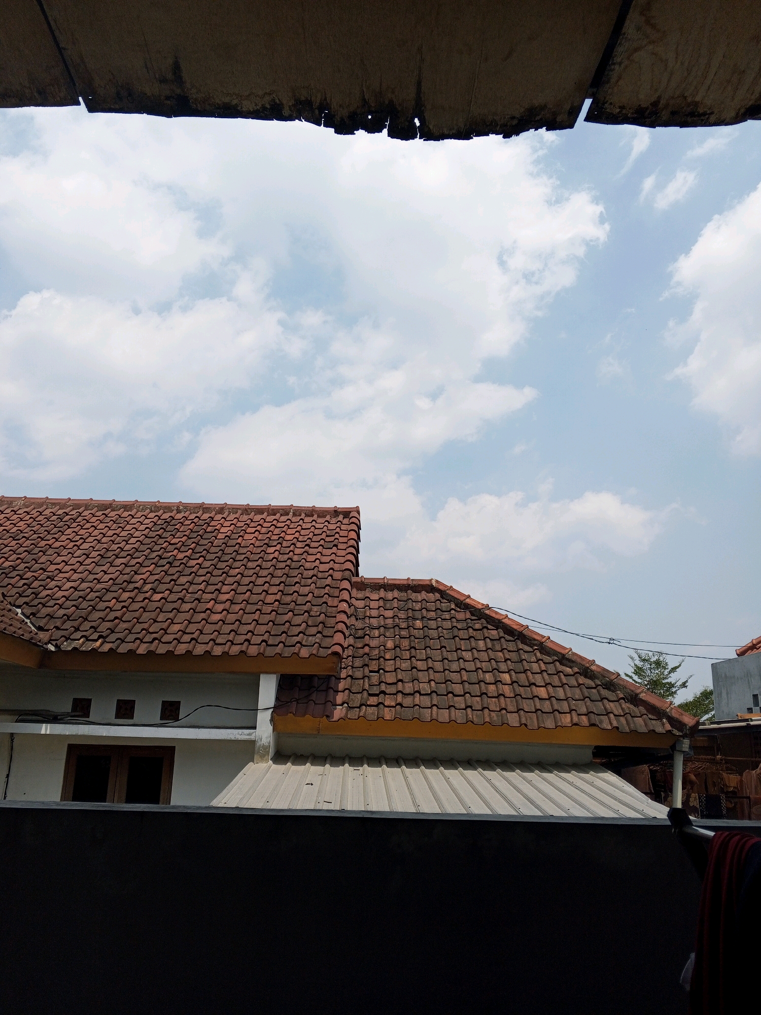The image shows a view of rooftops in Condongcatur, Indonesia, under a bright, cloudy sky. In the foreground, a dark, solid wall forms the bottom of the frame, with a darker, textured material, possibly a tarp or awning, extending from the top edge of the image downwards. Behind the wall, two sections of tiled rooftops are visible, both covered in terracotta-colored, curved tiles. The roof on the left is part of a white building with a single, dark-paned window and three small, square vents above it. This roof has a yellow trim along its edge. To the right of this, a second, larger roof section extends outwards, also with terracotta tiles and yellow trim. Below this second roof is a lighter-colored, corrugated metal roof. The sky above is a clear blue, punctuated by scattered white cumulus clouds. Further in the background, a green tree is visible, along with parts of other buildings and a glimpse of laundry hanging on a clothesline to the far right. The scene appears to be an outdoor, residential area in the daytime.