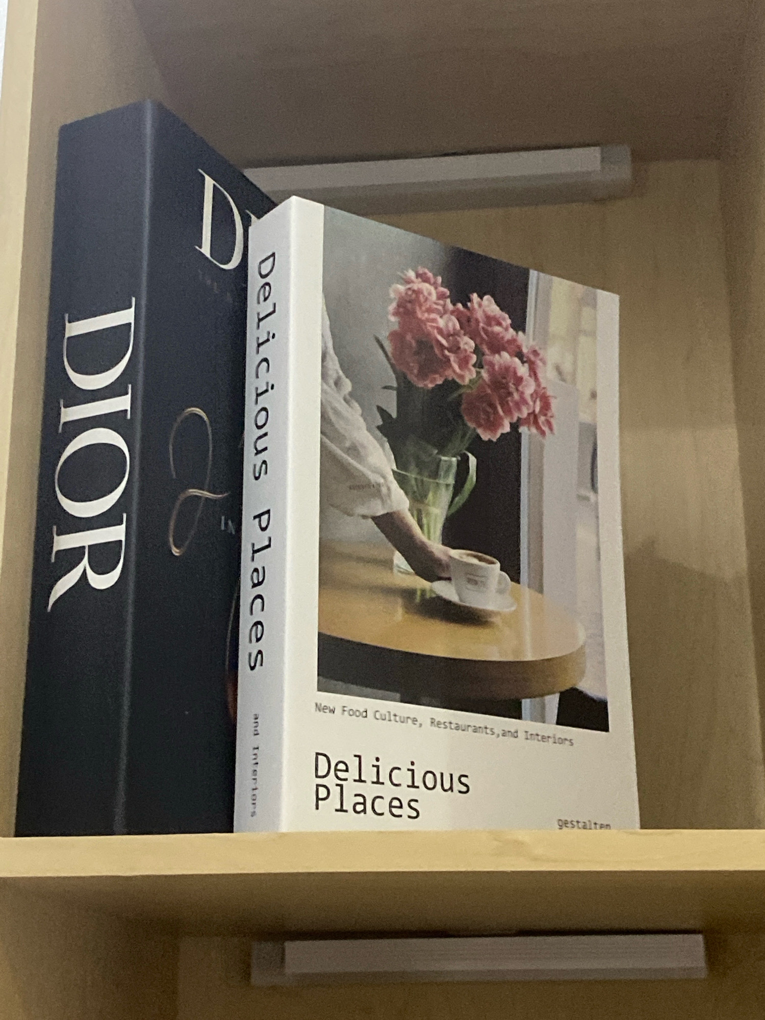 The image shows two books on a wooden shelf. The book on the left is black with "DIOR" in large white letters and a golden cursive "L" with "IN" below it. The book on the right is white with a picture of pink flowers in a vase on a table with a hand reaching for a cup of coffee. The spine of the white book reads "Delicuous Places". The lower portion of the white book has the text "New Food Culture, Restaurants, and Interiors" and "Delicous Places" with "gestalten" below. There is a white light fixture above the books. The scene appears to be an indoor setting, possibly a home or a bookstore, with wooden shelving and a neutral-colored wall. There is no specific indication of the location being in Kota Dalam, Indonesia.