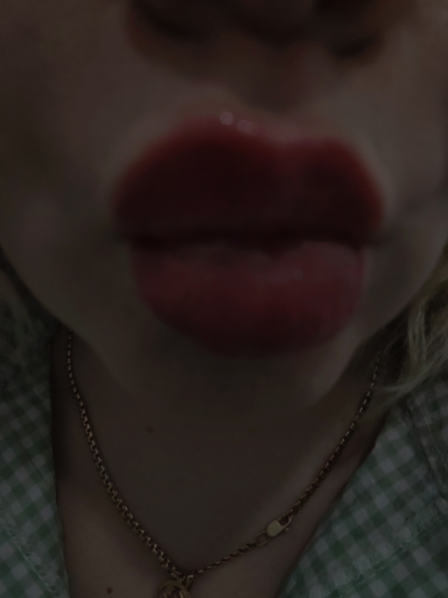 A close-up image focuses primarily on a person's face, specifically the lips. The lips are full and coated with a glossy, red lipstick. Above the lips, the blurry suggestion of a nose is visible. Below the lips, the image includes a portion of the person's neck. A gold necklace with a pendant hangs around the neck. The person is wearing a garment with a green and white checkered pattern. The background is generally dark and blurry. There is no information to determine that this image was taken in Izhevsk, Russia, and I cannot generate information about location if it is not provided in the source.