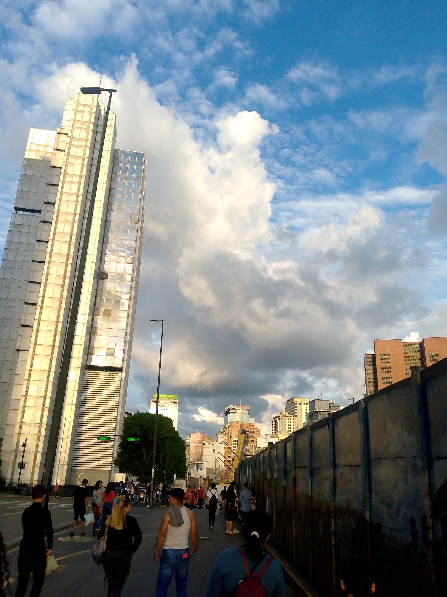 The image shows a street scene in Caracas, Venezuela. In the background, tall modern buildings reflect sunlight against a backdrop of blue sky and large, white cumulus clouds. A construction barrier extends along the right side of the frame, obscuring parts of the buildings behind it. Several people are walking in the street, some appearing to cross the road. Their clothing includes casual attire like jeans, t-shirts, and bags. The lighting suggests it is late afternoon or early evening. The image captures an ordinary urban moment amidst the city's infrastructure.