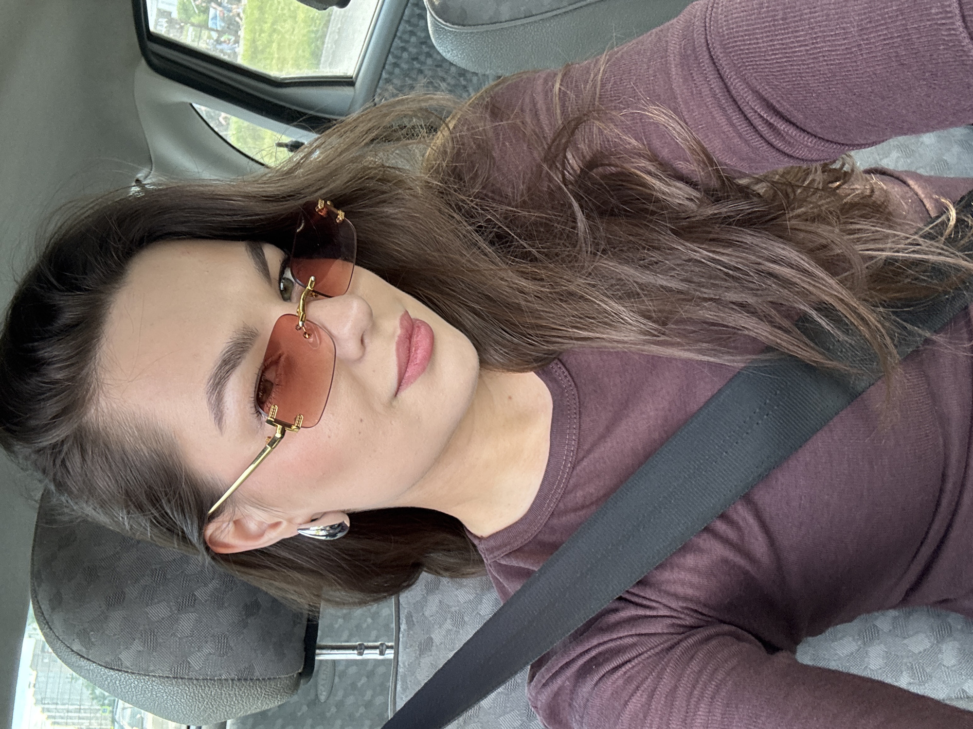 The media file shows a young woman in a car, likely in Chaikovsky, Russia. She is wearing a burgundy long-sleeved shirt and a black seatbelt across her chest. The woman has long, brown hair, light skin, and is wearing distinctive rimless sunglasses with a golden frame and gradient brown lenses. She also has large, silver hoop earrings. The background shows the interior of a car, including a headrest and a window with a blurred view of greenery and some buildings outside, suggesting daytime. Her facial expression is neutral, with her lips slightly parted. The image is a selfie, taken from a slightly elevated angle, capturing the woman from the chest up, with her head turned to the side. There is no visible text in the image.
