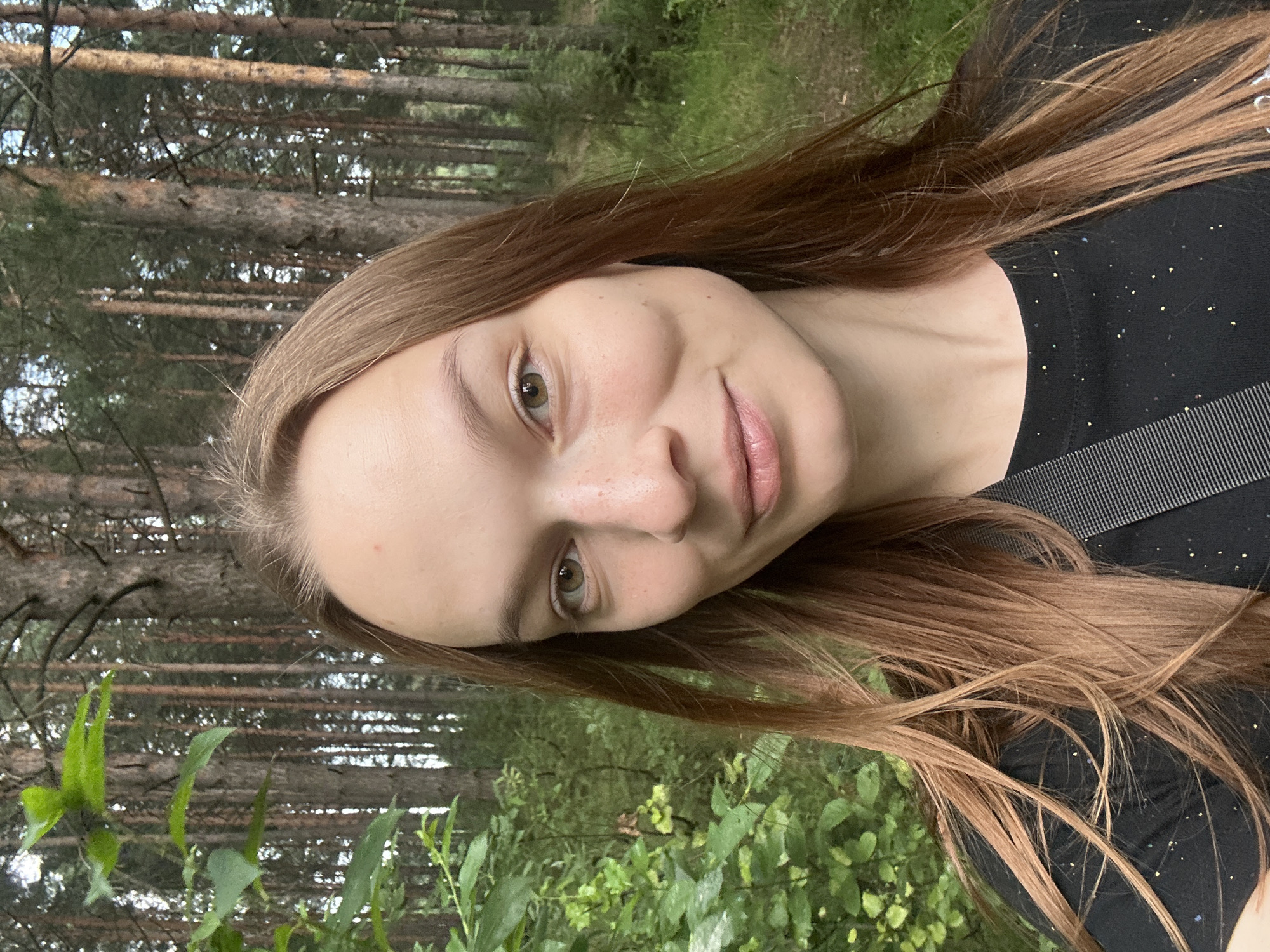 The image is a portrait of a young woman with long, light brown hair. She is wearing a black t-shirt with a subtle sparkly pattern and a strap visible over her shoulder. Her face is turned towards the camera, with her head tilted. She has light skin with freckles, green eyes, and light pink lips. A faint smile is visible. The background consists of a forest with tall trees and lush green foliage. The lighting suggests it might be daytime, possibly overcast due to the soft light. The image appears to be taken outdoors in a natural setting. There is no visible text in the image.