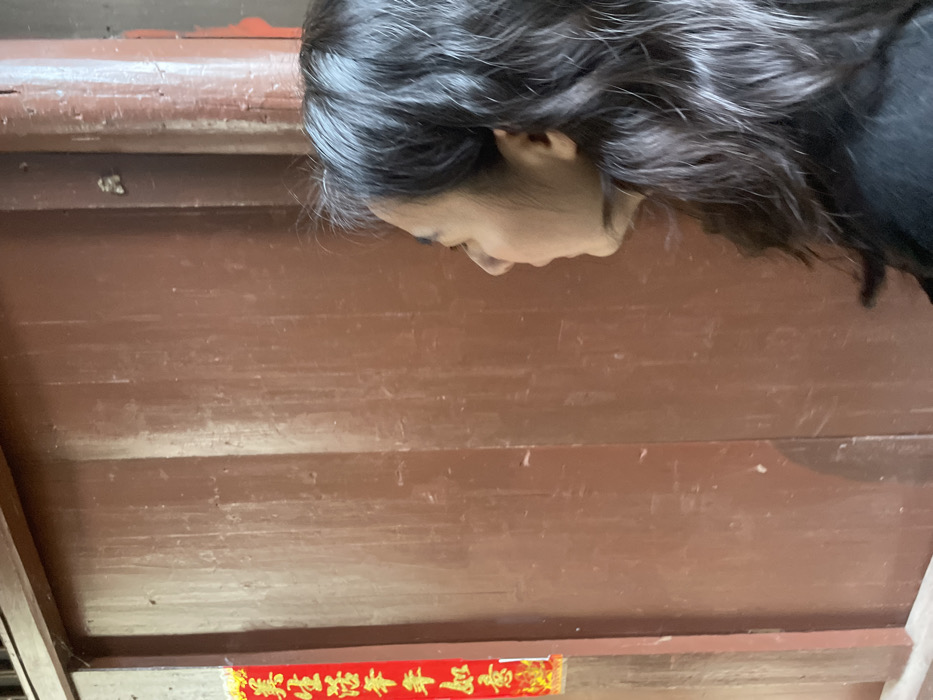 A woman with dark hair bends over a brown, wooden surface. The surface has horizontal planks, and a red and gold decorative banner is visible at the bottom. The woman's head is angled downwards, obscuring her eyes but showing her nose, mouth, and ear. Her hair is thick and slightly wavy. She appears to be looking at something on or below the wooden surface.