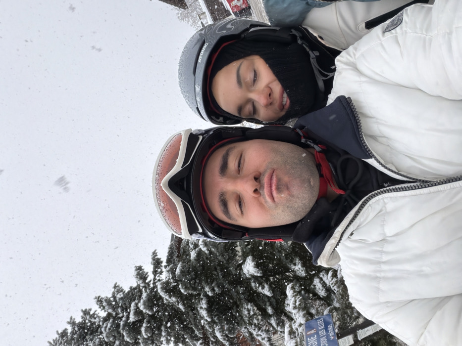 A close-up selfie depicts two individuals outdoors during active snowfall, confirmed by a visible sign indicating "SECTION LES ALLUES" and "MÉRIBEL" in France.
In the foreground on the left, a male subject wears a black ski helmet with white-framed goggles resting on top, a black balaclava or neck gaiter, a black hooded sweatshirt, and a white puffer ski jacket. He has visible stubble and is looking directly at the camera with his lips puckered.
Behind and to his right, a female subject wears a dark grey ski helmet with "Atomic" branding, a black balaclava covering most of her face, and a light blue/teal and white ski jacket. Her eyes are partially closed, and her head is slightly tilted towards the male.
The background features a heavily snow-covered evergreen tree on the left and a partially visible building or chalet on the right. The aforementioned blue sign is located on the lower left, partially obscured by snow. Numerous snowflakes are visible in motion across the frame, indicating ongoing precipitation. The environment is uniformly white and diffuse due to the snow and overcast conditions.