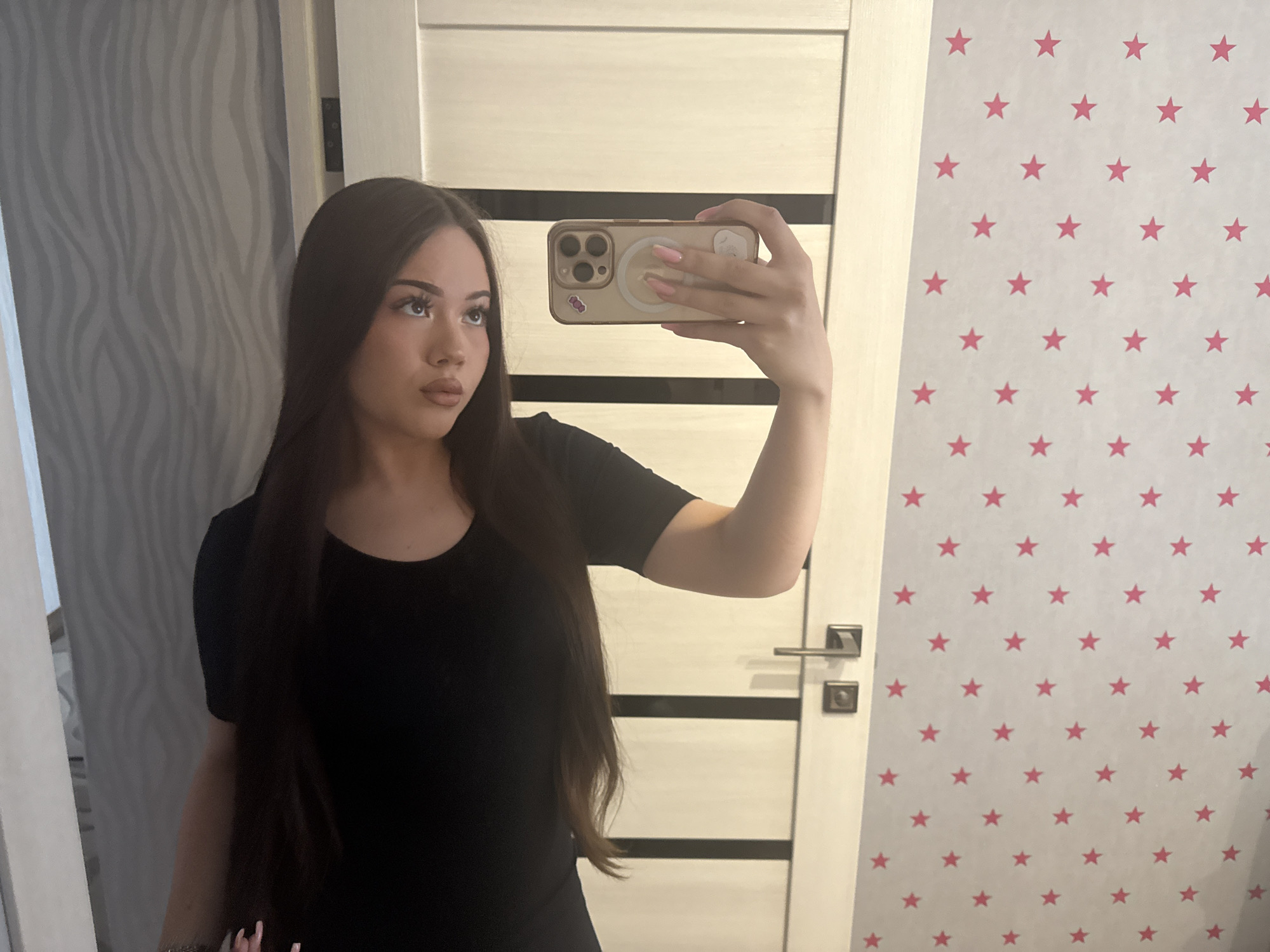 A young woman, likely in her late teens or early twenties, is captured in a mirror selfie in an indoor setting. She has long, dark brown hair that falls over her shoulders and wears a simple black short-sleeved top. Her gaze is directed slightly upwards and to her right (viewer's left), with a neutral expression. She holds a light-colored smartphone with three camera lenses in her right hand, positioned to take the photo. The phone case is transparent with a circular magnetic (MagSafe) attachment on the back and a small sticker, possibly a cartoon character, near the bottom. Her fingernails are long and manicured with a light pink or nude polish.
The background, visible as a reflection, features a light-colored door with dark horizontal panels or stripes directly behind her. The door has a modern-style handle and lock. To the left of the door (viewer's left), a wall with a gray, wavy or abstract patterned wallpaper is visible. To the right of the door, another wall is covered with white wallpaper featuring a repeating pattern of small, bright pink stars. The lighting appears to be ambient indoor light. The overall scene suggests a private room in a home located in Naberezhnye Chelny, Russia. No discernible text is visible in the image beyond what might be on the small phone sticker.