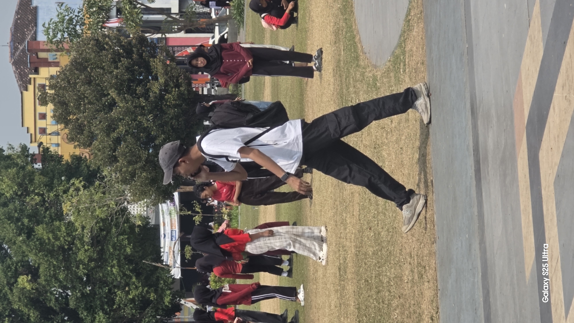 A man wearing a white t-shirt, black cargo pants, a gray baseball cap, and athletic shoes is walking and talking on a mobile phone in the foreground. He carries a black backpack. He is on a paved walkway bordering a grassy area in a park. In the background, several young women in red shirts and hijabs are standing and walking on the grass. Some are wearing white pants or plaid pants. Trees and buildings, including a yellow structure with a red roof, are visible behind the people. The overall setting appears to be an outdoor public space in a city.