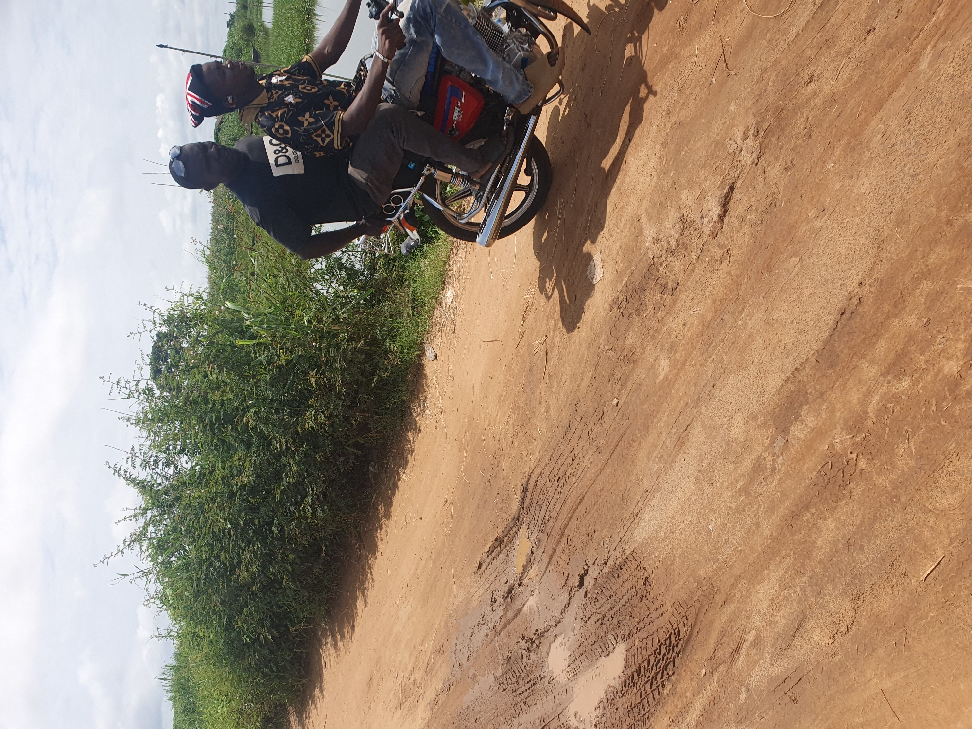 Two men are depicted on a motorcycle on a dirt road in Idah, Nigeria, under a cloudy sky. The image is taken from a slightly low angle, showing the road, lush green vegetation, and the sky.
The man driving is wearing a black t-shirt with "D&G" prominently displayed, and "DOLCE&GABBANA" in smaller text beneath it. He also wears sunglasses. The passenger behind him is dressed in a patterned shirt, possibly with a luxury brand motif, and a distinctive red, white, and blue patterned cap or headwear. The men appear to be mid-journey, possibly paused or moving slowly along the unpaved path.
The motorcycle is a standard model, with visible chrome exhaust pipes, wheels, and parts of its engine and frame. A red section, likely the fuel tank, is partially visible. The shadows cast on the ground suggest it is daytime, with the sun obscured by clouds, leading to softer lighting.
The setting is an outdoor, possibly rural or semi-rural area. The road is made of reddish-brown dirt, showing numerous tire tracks and some muddy puddles, indicating recent moisture. To the left side of the road, thick, tall green bushes and vegetation grow, hinting at a natural environment, potentially bordering a body of water visible in the distant background. The sky is largely overcast with white and grey clouds.