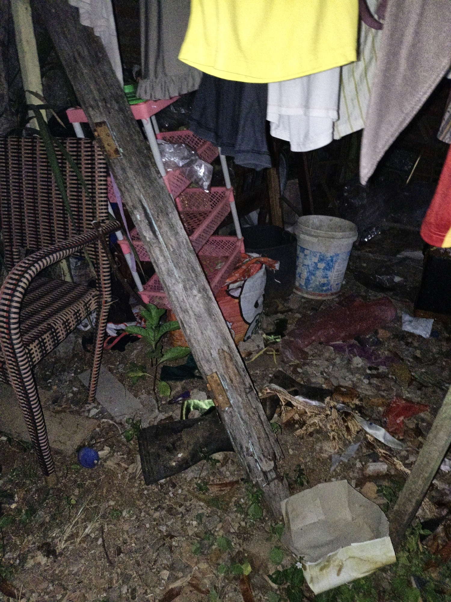 The image depicts an outdoor scene, possibly a backyard or alleyway, with a variety of objects scattered on the ground. A large, weathered wooden plank is a prominent feature, leaning diagonally across the frame. To the left, a wicker chair with a patterned seat and back is visible. Behind the chair and plank, a pink plastic shelving unit with multiple tiers is present. Various items of clothing are hanging in the background, including a yellow garment at the top. In the middle ground, a white plastic bucket with blue markings is situated on the ground. The ground itself is covered with debris, including leaves, dirt, and what appear to be fabric scraps. A small green plant is growing near the base of the wooden plank. In the foreground, a blue spherical object is on the ground, and a black rectangular object is also visible. The scene appears to be dimly lit, suggesting it was taken at night or in a poorly lit area. The location is identified as Cikal, Indonesia.