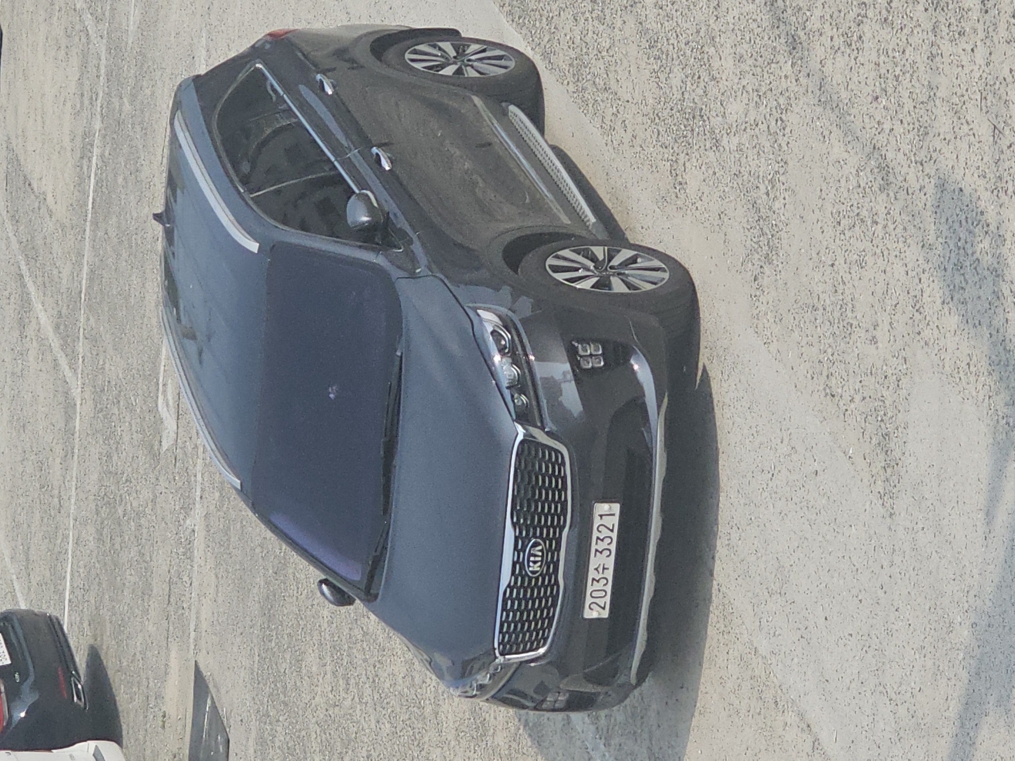 A dark gray Kia Sorento SUV is parked on a paved surface, likely a parking lot, in Hwaseong-si, South Korea. The vehicle has a license plate that reads "203 3321". The SUV has silver roof rails and chrome accents on the door handles and side steps. The front grille features the Kia logo. In the background, the rear of another vehicle, possibly a black sedan, is visible. The ground is a rough, light-colored asphalt with visible gravel. The lighting suggests it is daytime.