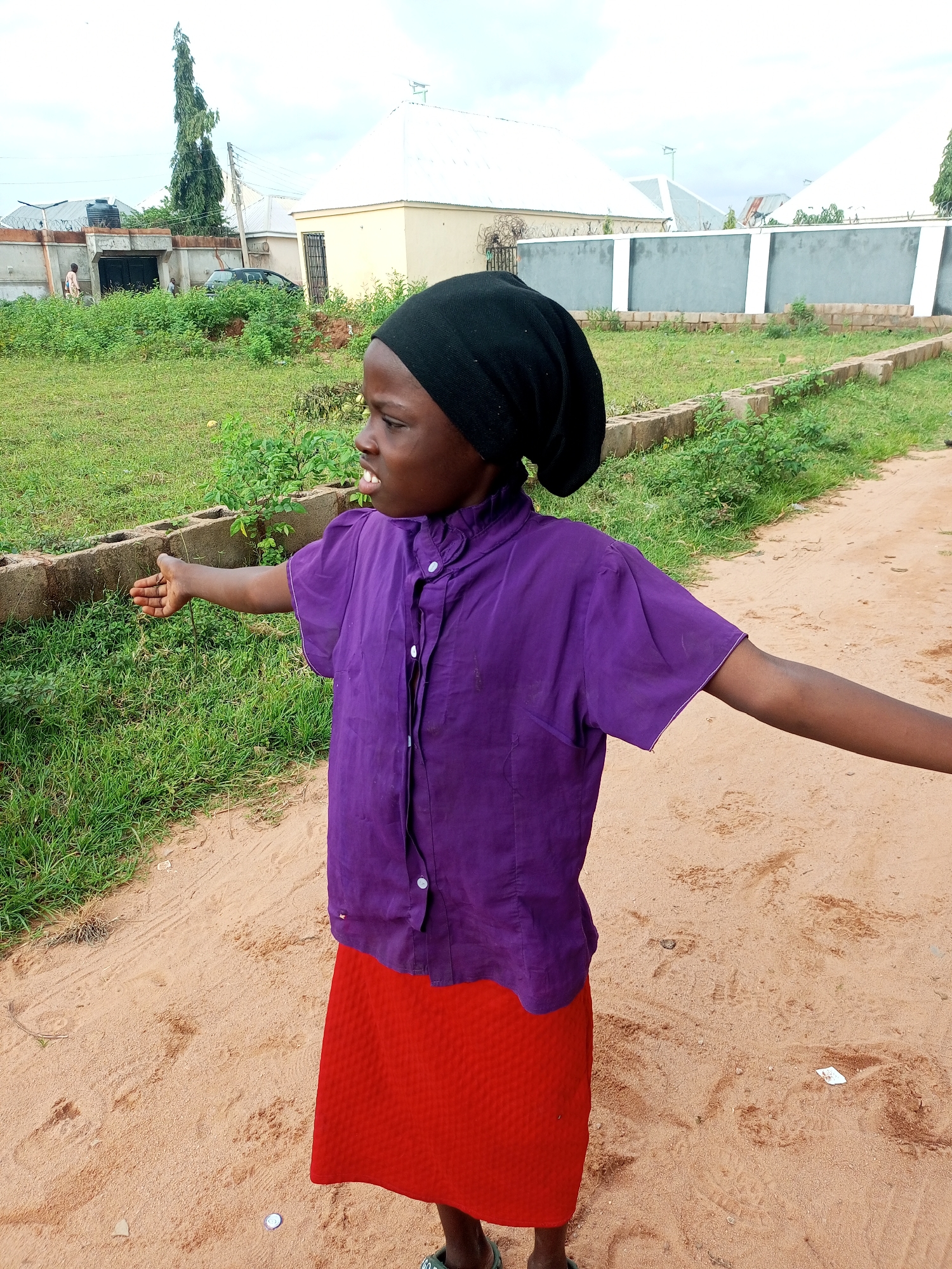 A young girl wearing a black headscarf, a purple short-sleeved shirt, and a red skirt stands on a dirt path. Her left arm is extended to the side, with her palm open. In the background, there is lush green grass, a low brick wall, and a grey wall with white accents. Beyond the walls are several buildings with white roofs, and a tall, slender tree. The scene appears to be an outdoor setting in the city of Tempure, Nigeria.