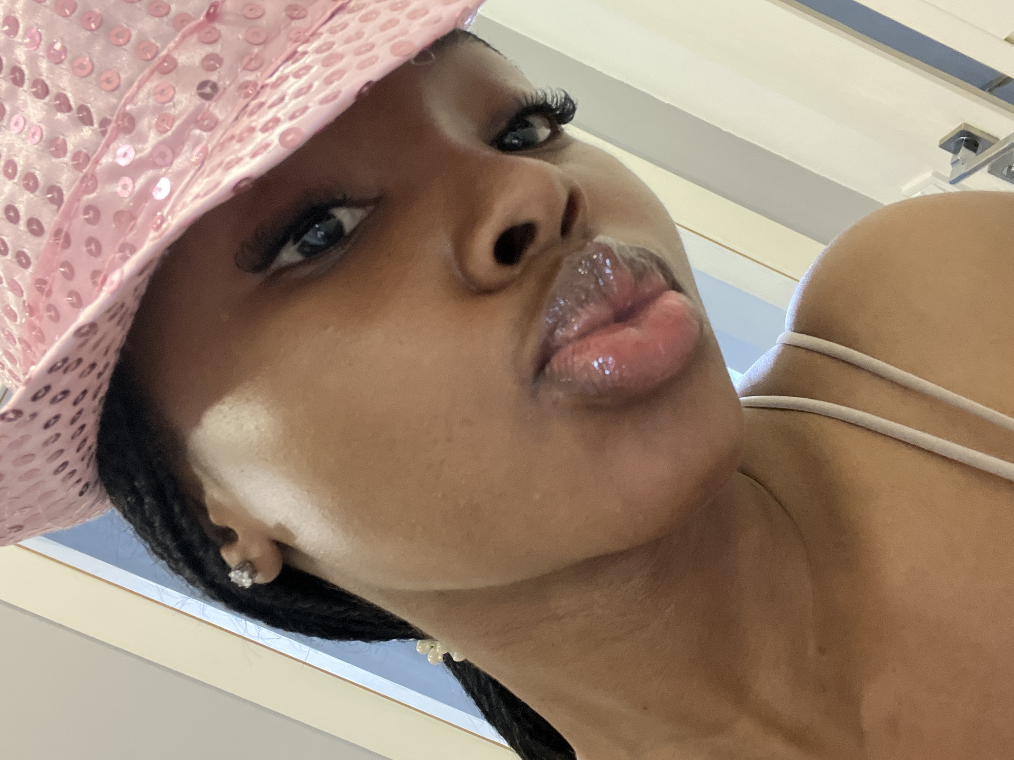 A close-up, tilted, selfie-style shot shows a young Black woman wearing a pink sequined hat and a white tank top. She has braided hair, which is partially visible, and is wearing a diamond stud earring. Her lips are glossy and pursed. The background is out of focus and appears to be indoors, with a door and wall visible. The image does not contain any identifiable landmarks or clues suggesting it was taken in Kazan, Russia.