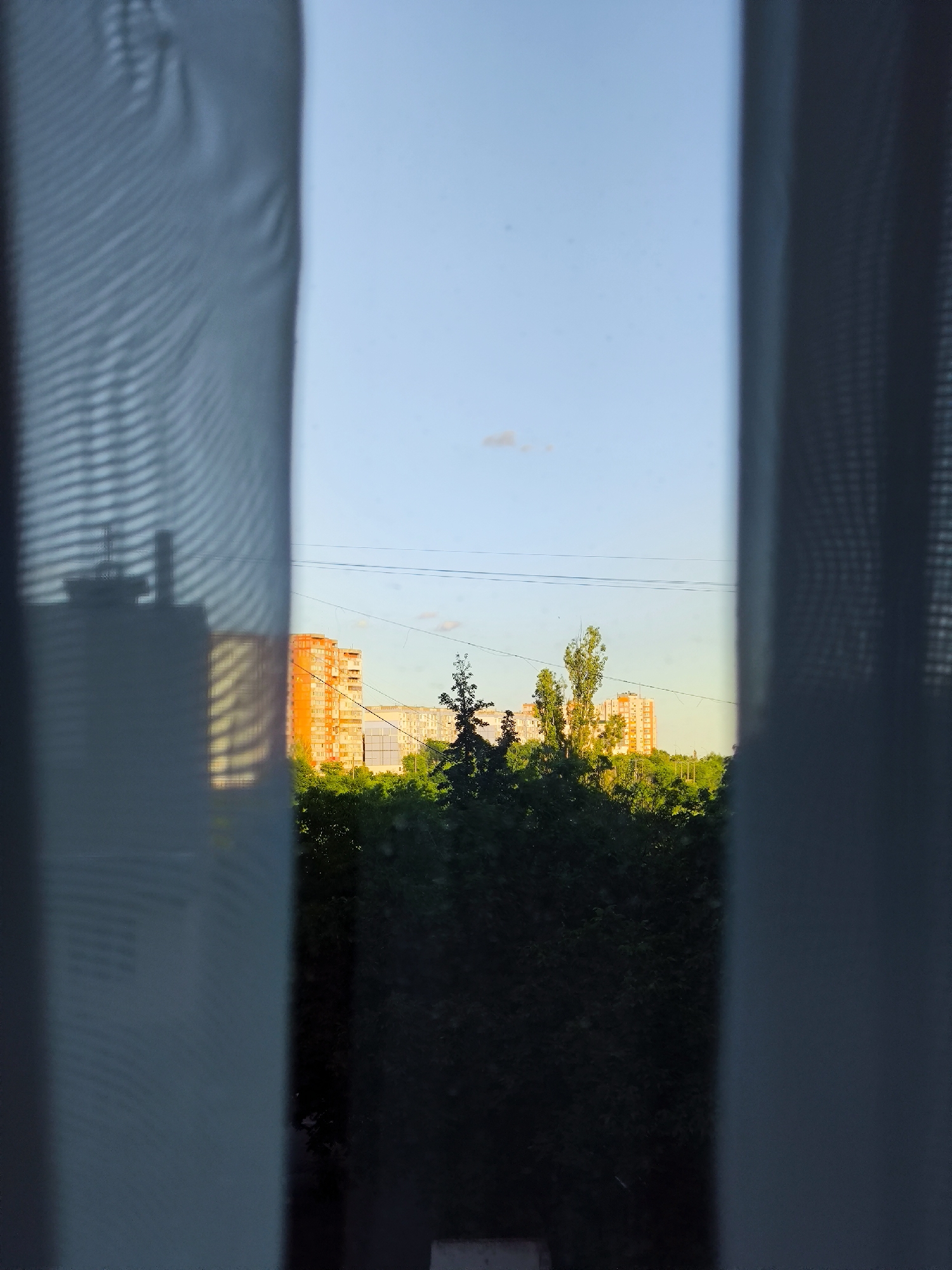 The image captures a view through sheer curtains, revealing a residential scene at dusk. In the foreground, the soft folds of white and gray sheer curtains frame the view, with the left curtain showing a textured pattern and casting faint shadows. The main subject is an apartment building complex bathed in the warm, golden light of the setting sun. The buildings are primarily orange and white, with numerous windows. They are nestled amongst lush green trees, suggesting a summer or early autumn setting. A few tall, slender trees stand out against the backdrop of the buildings and the sky. The sky is a clear, pale blue, with a hint of a pale cloud near the horizon, indicating the transition from day to evening. Overhead, thin power lines are visible, a common sight in urban or suburban environments. The overall mood is serene and peaceful, conveying a sense of quiet observation from indoors looking out at the urban landscape. There is no visible text in the image.