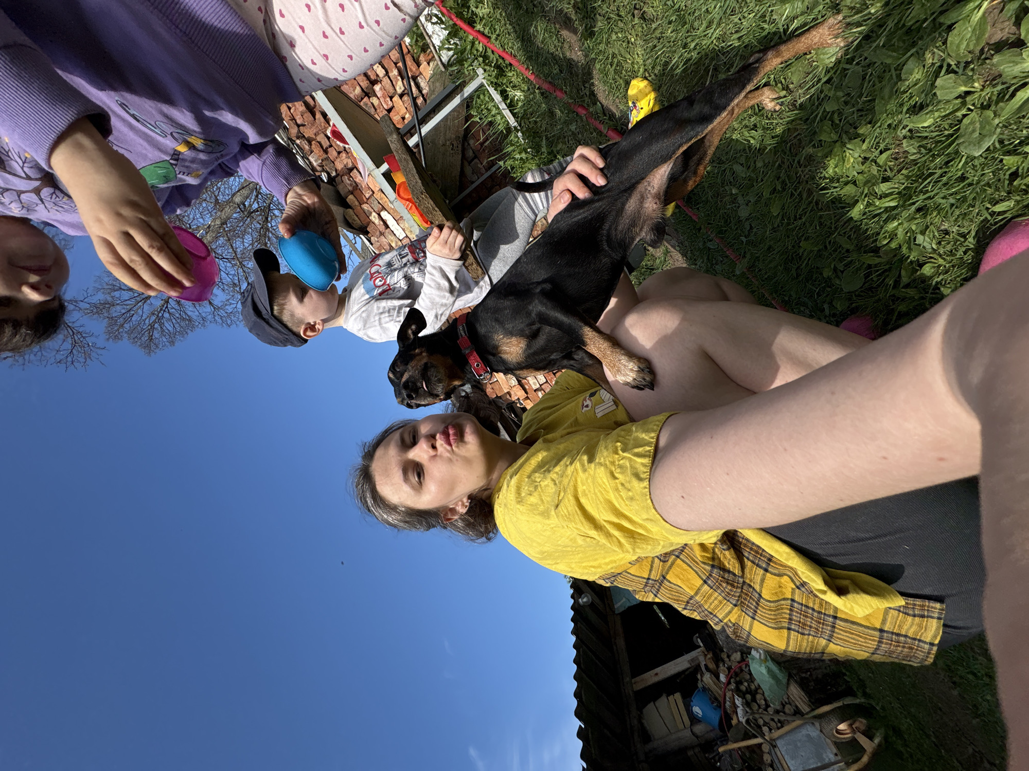 The image shows a group of people and a dog outdoors. A woman in a yellow shirt and plaid vest is holding a black and tan dog on her lap. Two children are partially visible above, holding blue and pink plastic Easter eggs. Behind them is a stack of bricks. The sky is blue. The location cannot be verified with the available information.