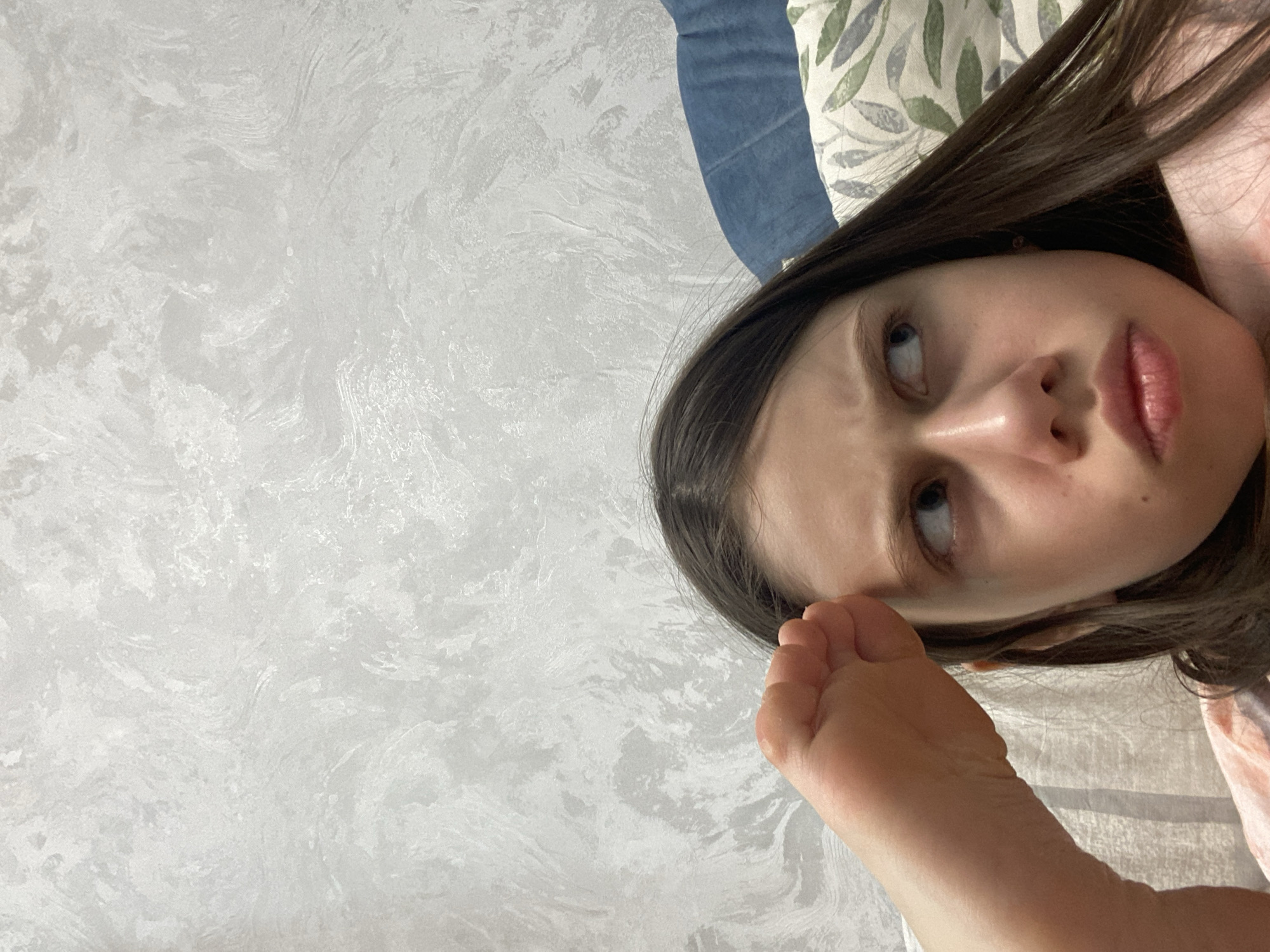 The image is a close-up selfie of a woman with fair skin, brown hair, and blue eyes. Her face is tilted, and she is looking upwards. Her right hand is raised near her face. The background consists of a light grey textured wall, a partially visible blue pillow, and a pillow with a leaf pattern. There is no information available in the image to indicate that the location is in the city of Izhevsk, Russia.