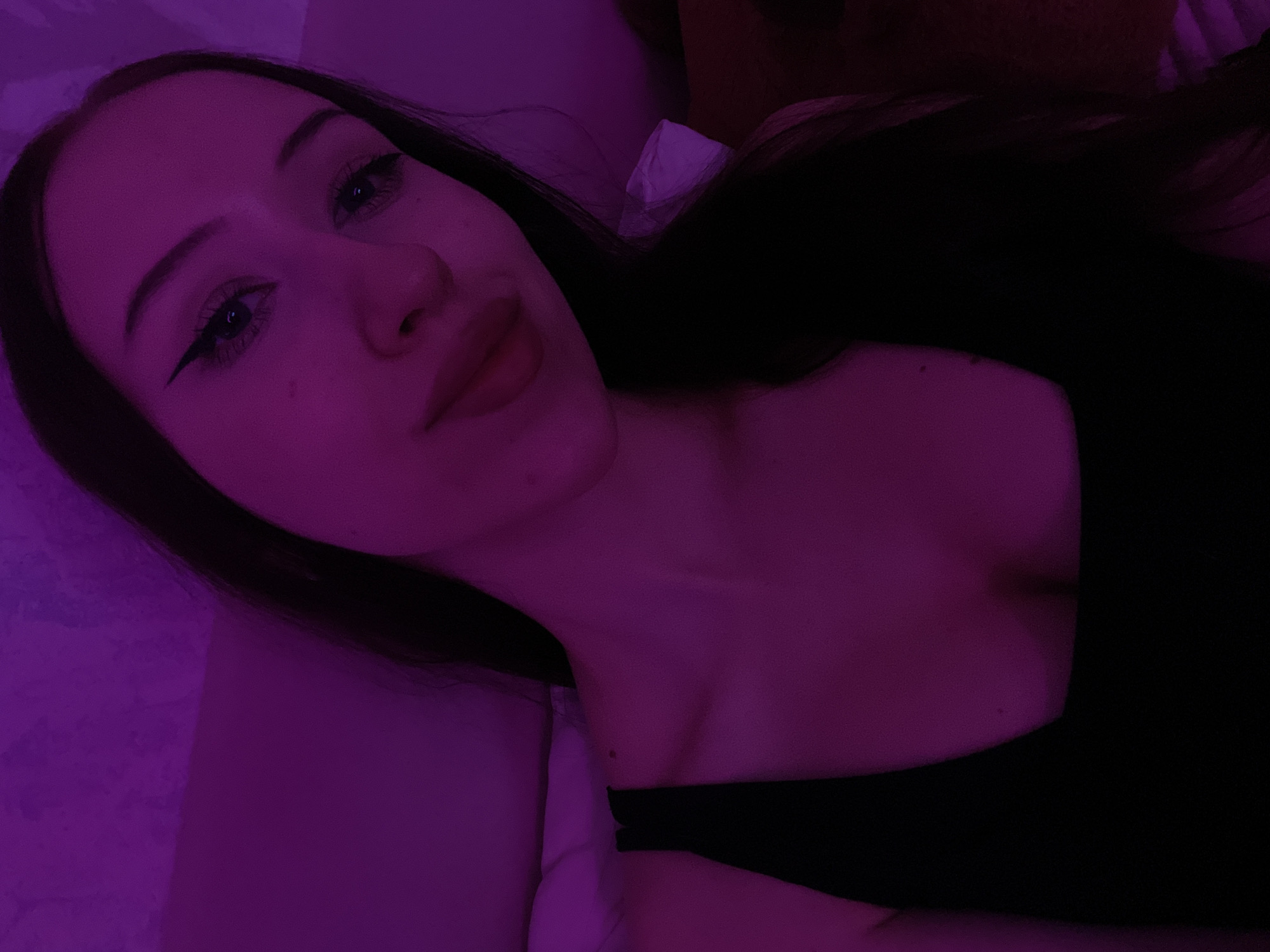 A young woman with long, dark hair is captured in a close-up selfie, illuminated by purple light. She is wearing a black top. Her makeup includes winged eyeliner and lipstick. The background appears to be a textured wall. The image was taken indoors, likely in a bedroom setting. No specific location in Votkinsk, Russia, is discernible from the visual information.