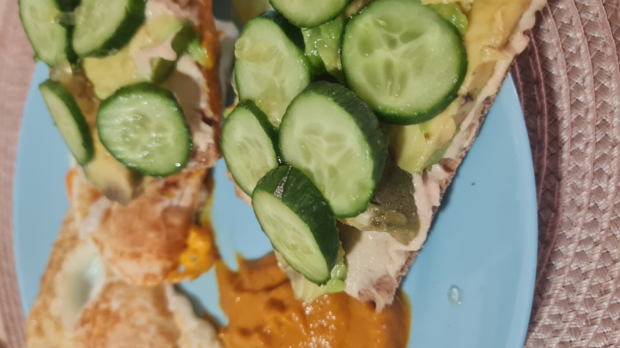 A close-up shot on a light blue plate displays a meal composed of two open-faced sandwiches topped with sliced cucumbers, accompanied by fried eggs and what appears to be a sauce, possibly hummus or another spread, on the side. The sandwiches seem to be on whole-wheat bread and are spread with a creamy substance. Avocado chunks are interspersed with the cucumber slices on the sandwich. The surface beneath the plate is textured with a tightly woven pattern. There is no indication of the location being Nahariyya, Israel or any indication of people in the image.