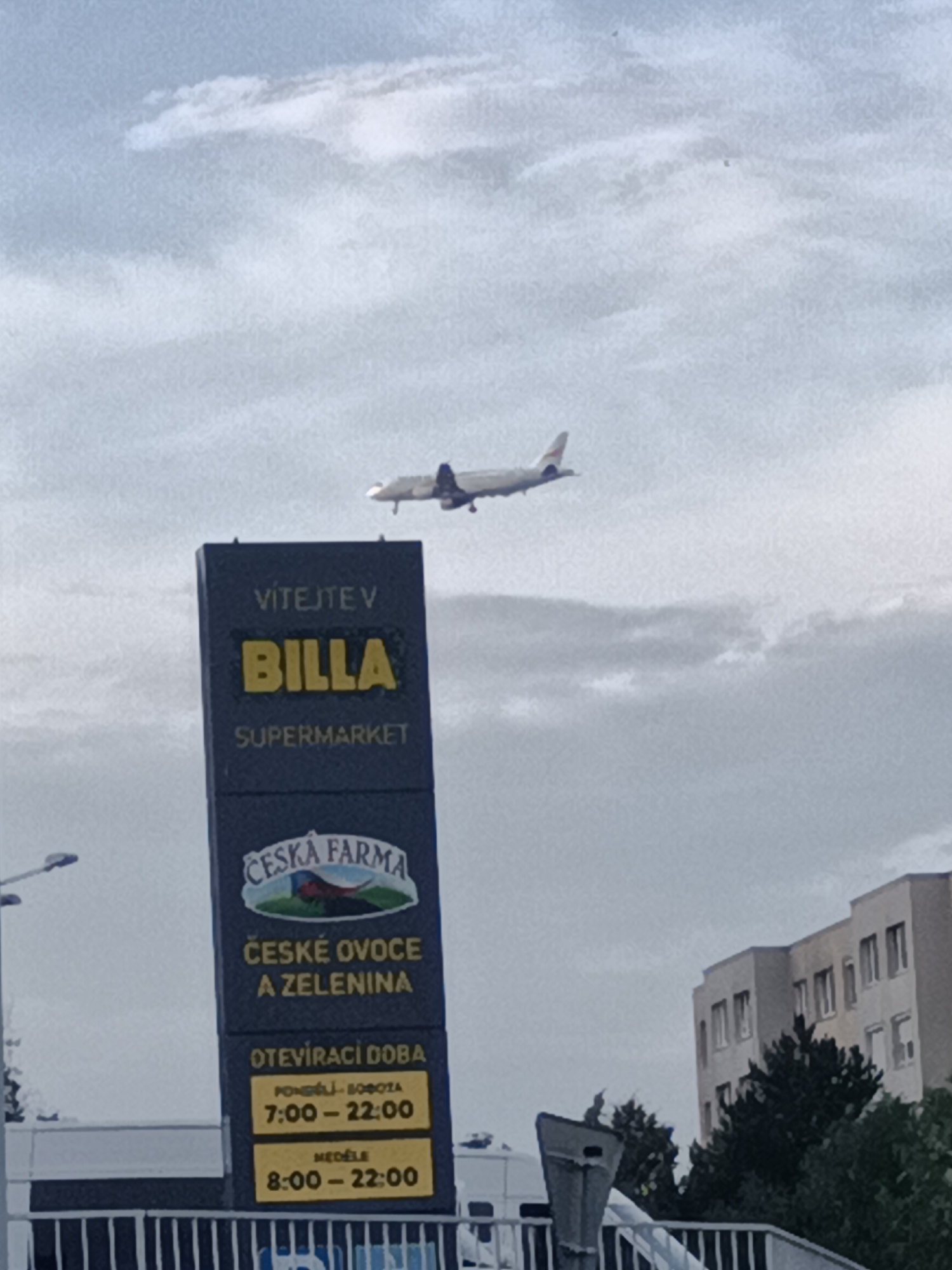 The image captures an airplane in mid-flight against a cloudy sky, positioned above a prominent dark blue sign. The sign displays "VÍTEJTE V BILLA SUPERMARKET" in white text, with "BILLA" highlighted in yellow. Below this, a logo for "ČESKÁ FARMA" is visible, followed by text indicating "ČESKÉ OVOECE A ZELENINA" (Czech fruits and vegetables). Further down, opening hours are listed: "PONDĚLÍ - SOBOTA 7:00 - 22:00" and "NEDĚLE 8:00 - 22:00". To the right of the sign, a modern building complex with multiple windows is visible, along with lush green trees. The scene appears to be a commercial area, likely in Prague, Czechia, given the Czech language on the signage.