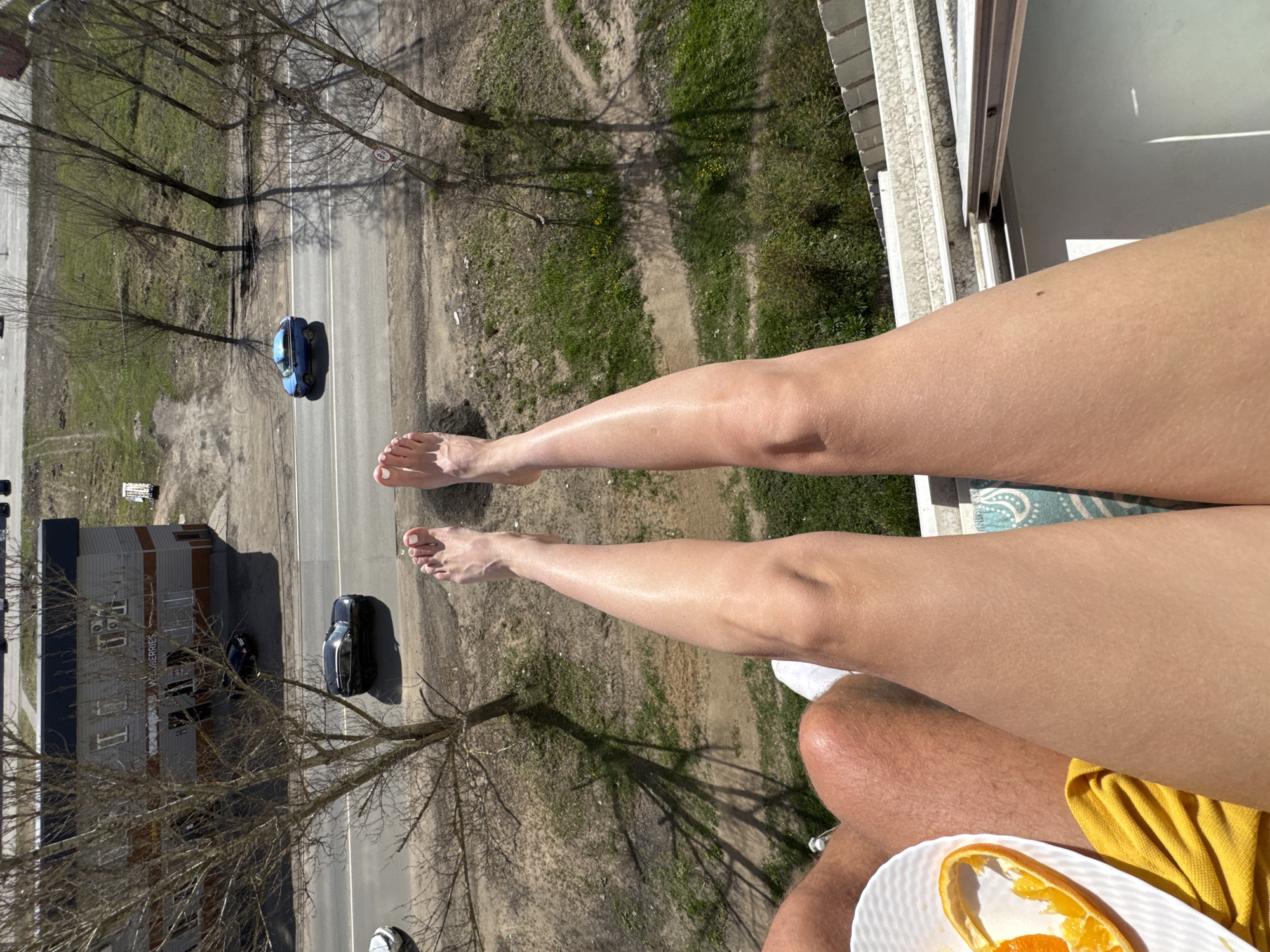 The image captures a top-down view from a balcony or high window in Ново-Никольское, Russia, showing two individuals relaxing while overlooking a street scene below.
In the foreground, a pair of bare, tanned legs, likely belonging to a female, are stretched out, with painted toenails visible on the feet. Adjacent to these, a portion of another person's leg and knee is seen, wearing white shorts, suggesting a male. A white, textured plate with orange peels rests near this person, beside a yellow cloth or garment. A blue patterned mat is partially visible beneath one of the legs, contributing to the relaxed atmosphere.
The view beyond the balcony railing reveals a residential or commercial street. Two cars are parked or driving on the asphalt road: a distinct blue sedan and a dark-colored car, possibly an SUV or another sedan. A wide grassy verge with scattered patches of dirt separates the road from a row of bare-branched trees, which cast long shadows, indicating a sunny day.
On the left side of the street, a multi-story building is visible, with a prominent sign on its side that clearly reads "WILDBERRIES," identifying it as a store or distribution center for the Russian e-commerce giant. Further down the street, another smaller building or structure is partially seen. A circular road sign with a red border and the number "80" inside, likely indicating an 80 km/h speed limit, is visible on a pole near the street.
The overall scene suggests a pleasant, sunny day, likely midday or early afternoon given the shadows, with people enjoying a leisurely moment outdoors on a balcony.