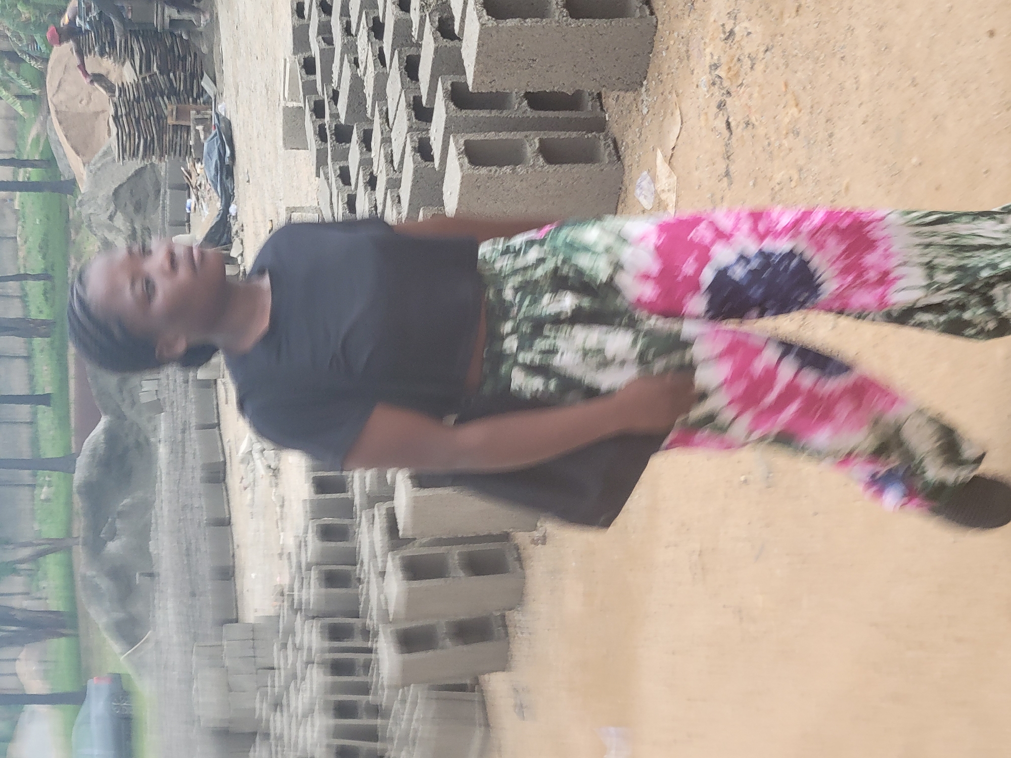 An outdoor, slightly blurry photograph, taken during the daytime in Kwamba, Nigeria, shows a young woman standing amidst a scene dominated by concrete blocks. The image is oriented vertically (originally rotated 90 degrees counter-clockwise).
The central subject is a woman, likely in her late teens or early twenties, with dark skin and braided or dreadlocked hair. She is wearing a plain black t-shirt and vibrant, tie-dye patterned trousers featuring shades of pink, green, white, and dark blue. A black bag is slung over her left shoulder. She is looking slightly to her left, with a neutral expression.
The ground she stands on is unpaved, appearing to be dirt or sand. To her right and behind her, numerous grey concrete blocks (also known as cinder blocks) are stacked in organized piles, suggesting a construction site or a block-making facility. In the mid-ground and background, more blocks are visible, along with a large mound of earth or sand. Faintly in the distance, another person can be seen working or moving near more stacks of materials and what appears to be part of a structure or machine. Green foliage and tree trunks are visible on the left side of the frame, indicating an outdoor, somewhat natural setting. A dark-colored water bottle is partially visible on the ground near the left edge.
The overall scene depicts an active work environment related to building materials or construction, with the woman perhaps observing or participating. No discernible text is visible in the image. The lighting suggests an overcast day or indirect sunlight.