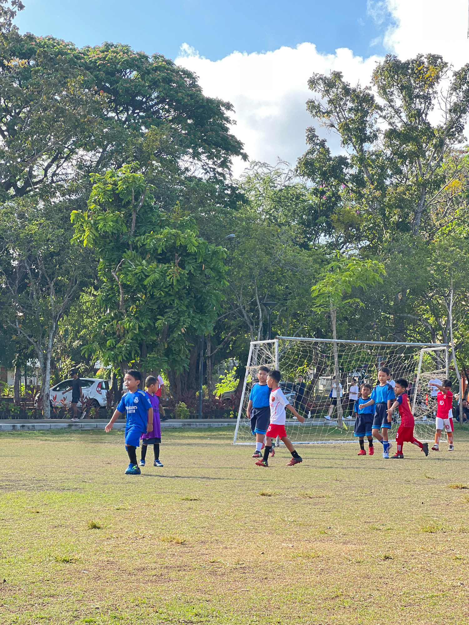 A group of young boys are playing soccer on a grassy field with a soccer goal in the background. The boys are wearing various colored jerseys and shorts. The field is surrounded by trees and there is a white car parked in the background. The sky is blue with some clouds. The scene appears to be taking place in Denpasar, Indonesia, as indicated by the lush greenery and tropical trees.