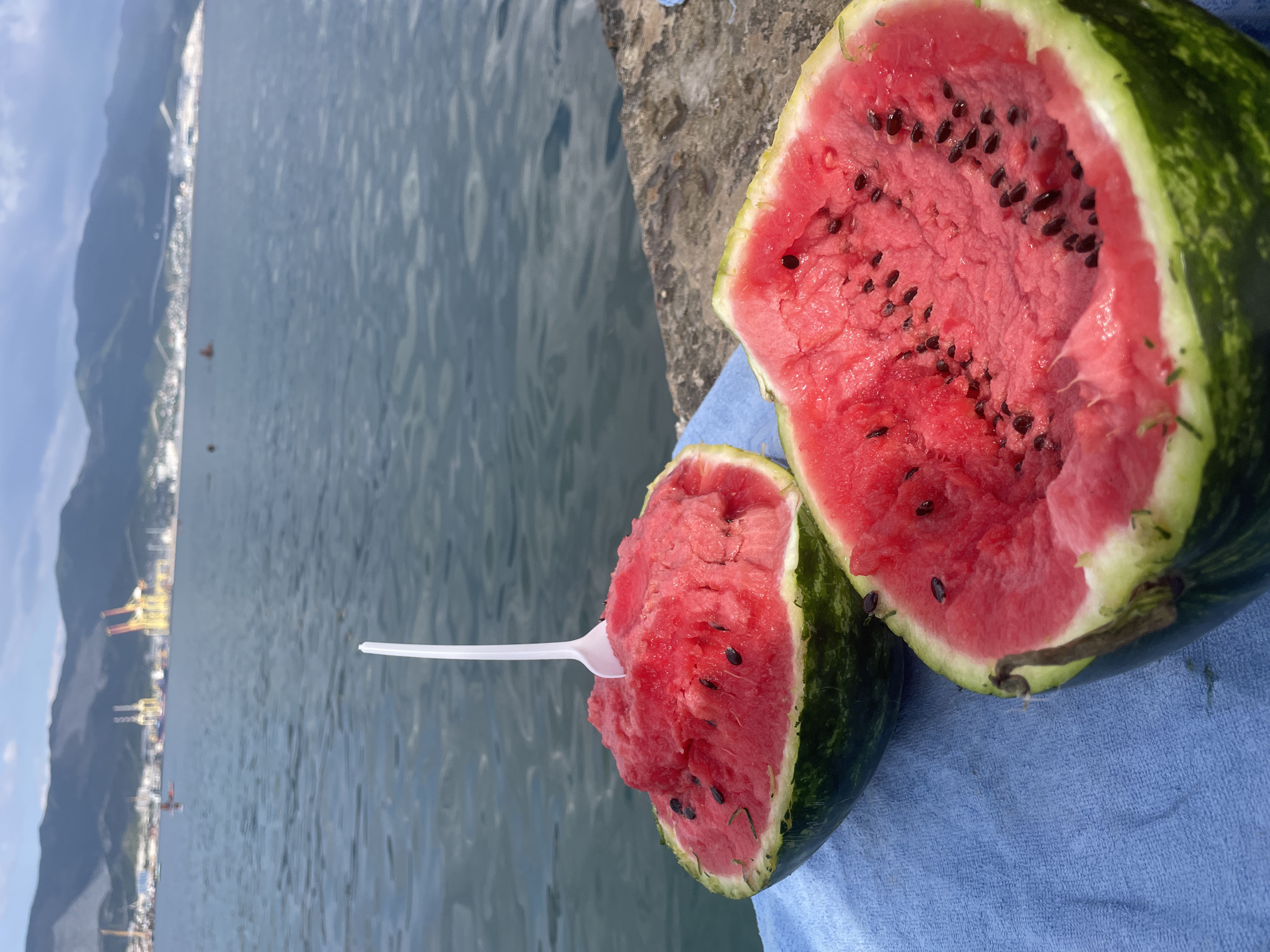 Two halves of a watermelon are resting on a blue towel next to the sea. One half of the watermelon has a white plastic spoon in it, with some of the watermelon scooped out. The other half is empty. In the background, there is a body of water, mountains, and some industrial cranes, suggesting the location is Novorossiysk, Russia.