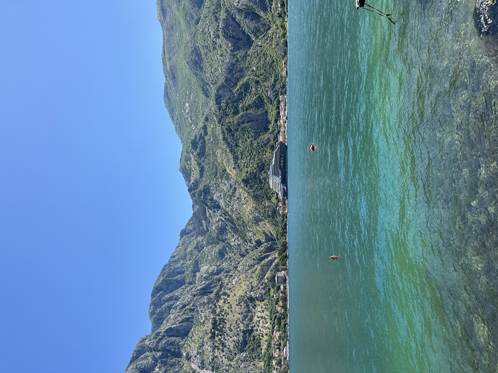 The photograph presents a view in Prčanj, Montenegro, featuring a mountainous landscape alongside the sea. The left side of the image showcases a rugged mountain range with dense vegetation. Nestled between the mountain and the shoreline are buildings, indicative of residential or commercial structures.
The right side depicts the sea, with varying shades of green and blue suggesting depth and differing light penetration. Two visible orange buoys float on the surface of the water. In the upper-right corner, there are fishing rods placed on the shore. The sky is clear blue, contrasting with the landscape below.