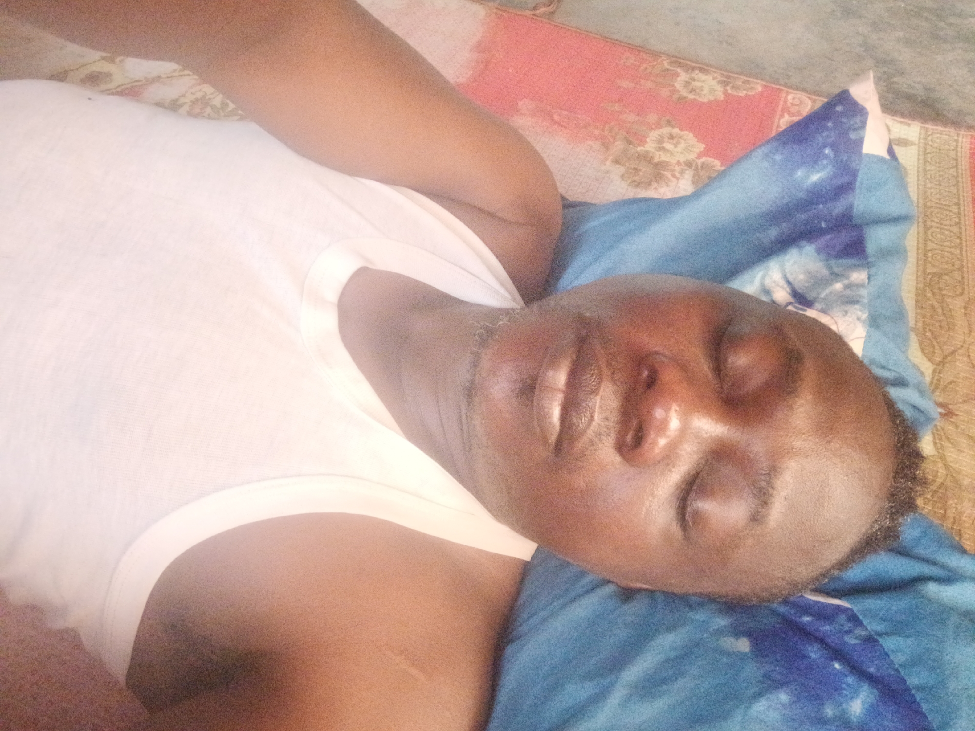 A person is lying down on a blue pillow. The person is wearing a white tank top. The background is a multi-colored carpet or rug. The location is unspecified.