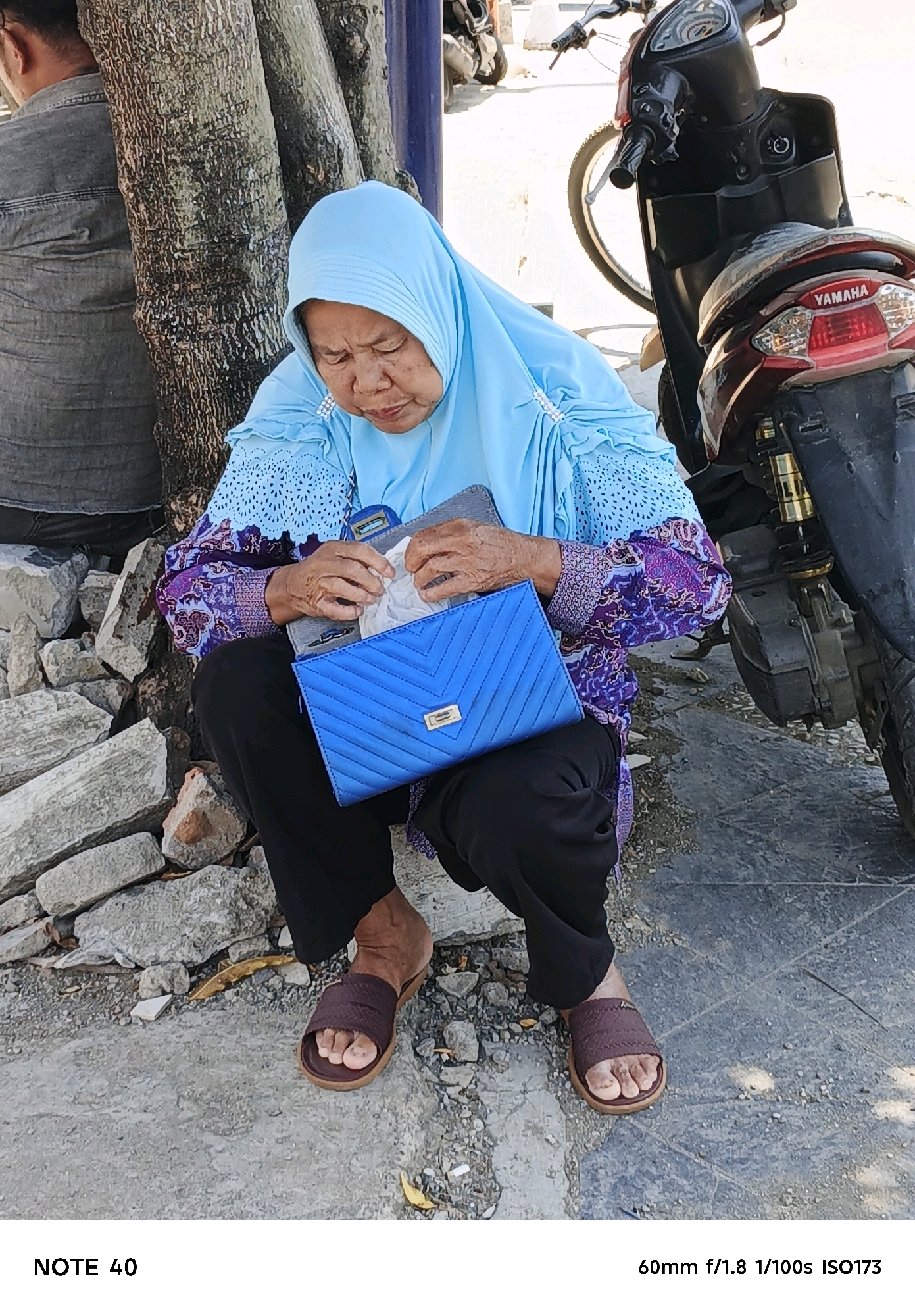 In the city of Kelurahan Sidanegara, Kecamatan Cilacap, Indonesia, an elderly woman wearing a light blue hijab and a vibrant purple and blue patterned top is seated on the ground, leaning against a tree trunk. She is holding an open, bright blue clutch purse and appears to be looking inside it. Black pants and brown slip-on sandals complete her attire. The ground beneath her is paved with a textured surface and scattered with small stones and dried leaves. A portion of a motorcycle, a Yamaha, is visible to the right of the woman, with its rear wheel and taillight prominent. To the left, another person, partially visible, is also leaning against the tree. The environment suggests an outdoor setting, possibly a roadside or public area.