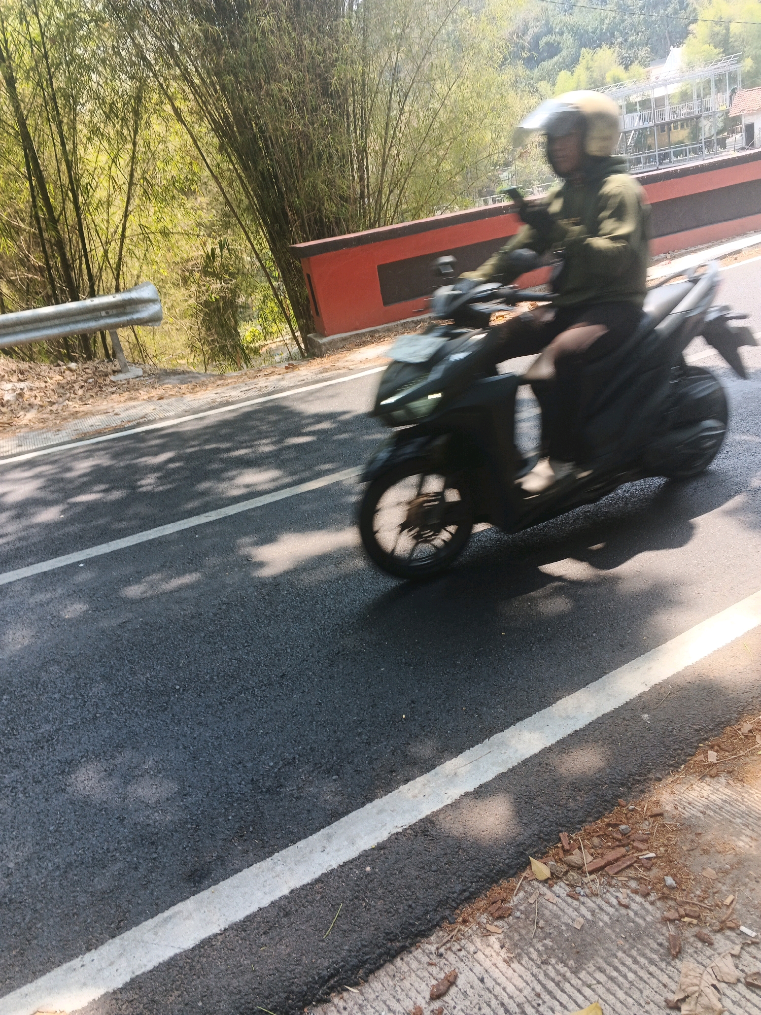 The image captures a person riding a scooter on a paved road with a white line marking the lane. The rider is wearing a helmet and a green jacket. To the left of the road, a metal guardrail is visible, and behind it, a dense bamboo forest covers a sloped area. On the right side of the road, there is a red barrier with a black stripe, and further in the background, buildings and more vegetation are visible. The scene is illuminated by sunlight, casting shadows on the road. The overall impression is of a rural or semi-urban environment, possibly on the outskirts of Semarang, Indonesia, given the presence of lush vegetation and buildings that could be found in such a region. The image is a still photograph, with the scooter in motion as indicated by the motion blur.