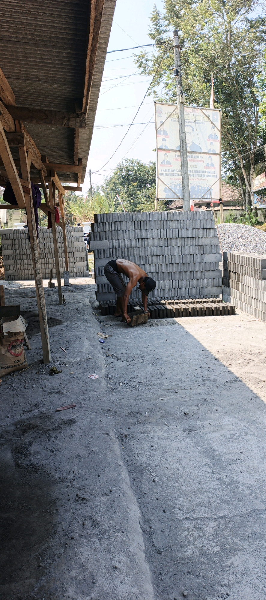 In Kasiyan, Indonesia, a shirtless person, wearing dark pants and a black cap, is bent over at the waist, manipulating a brick with both hands. They are positioned on a dirt ground surface. Behind them, a towering stack of grey bricks rises, with another smaller stack to their left. To the right, a pile of loose gravel is visible. The scene is partially covered by the corrugated metal roof of a simple wooden structure on the left, supported by wooden posts. A banner with portraits of two men and Indonesian text is affixed to a pole behind the brick stacks. Trees and a clear sky are visible in the background.