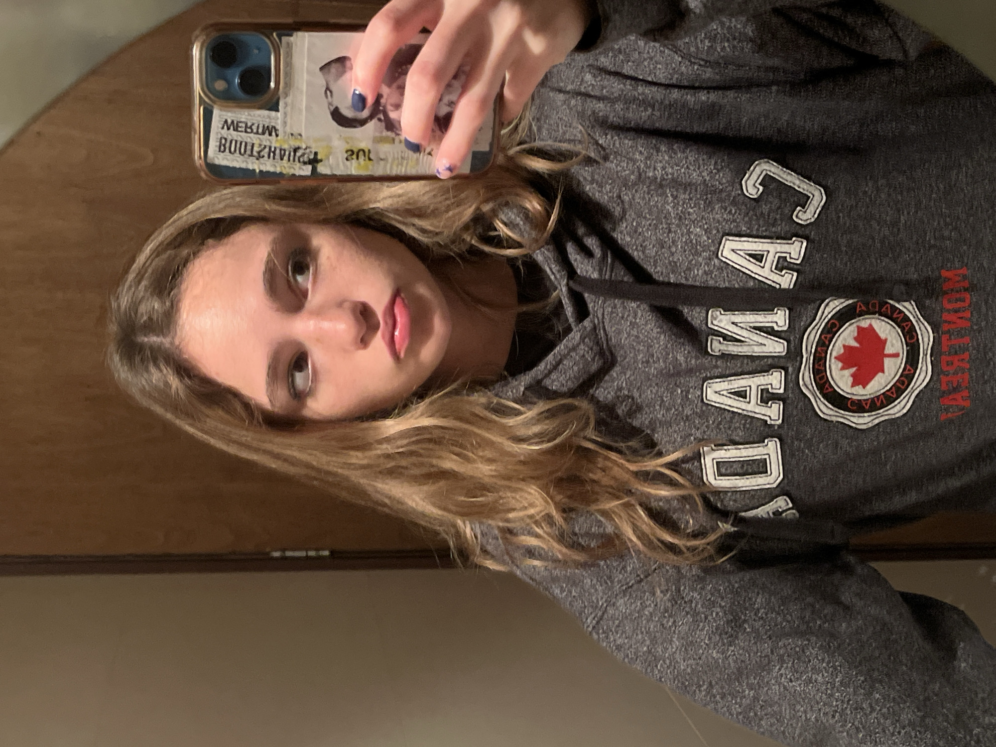 A young person is shown in a mirror selfie, holding a smartphone with their left hand. The subject has long, wavy blonde hair and is wearing a dark grey hooded sweatshirt. Their eyes look directly at the camera's reflection, with a neutral expression and slightly parted lips. The nails on the visible hand are painted dark blue.
The smartphone has a light blue case adorned with what appear to be multiple stickers or labels. Mirrored text visible on the case includes "MIMBUZHTYA" (likely reversed from "AYTHZUBNIM" or similar) and "20L".
The grey hoodie features large white, outlined letters spelling "CANADA" across the chest. Below this, there is a circular emblem containing a red maple leaf. Text around the emblem is also mirrored, reading "CANADA" along the top arc and "MONTRÉAL" along the bottom arc when viewed correctly, with other smaller, indistinct text. A dark drawstring is visible on the hoodie.
The background of the indoor setting includes a wooden surface, possibly a door or wall, and a lighter colored wall. The lighting appears to be artificial. The image is taken in San Bernardino Tlaxcalancingo, Mexico.