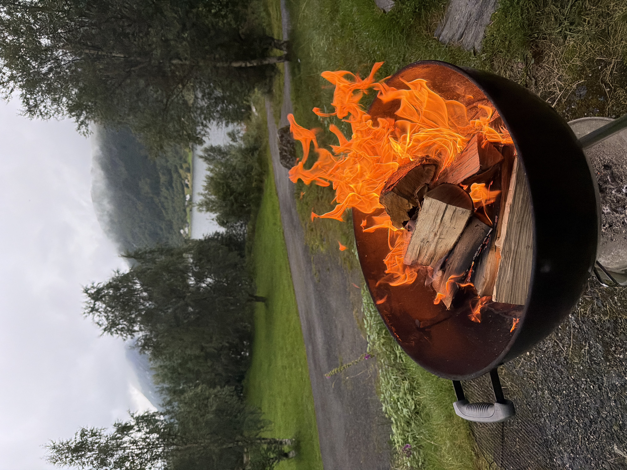 An outdoor barbecue with a fire burning is set up on a paved area next to a grassy lawn. The barbecue is a black kettle-style grill containing several logs of wood engulfed in bright orange flames. The grill has a black handle on the left side. In the background, there is a body of water, possibly a fjord, with tree-covered hills and mountains rising behind it. The sky is overcast and foggy, with the mist clinging to the tops of the mountains. The location is Skei, Norway.