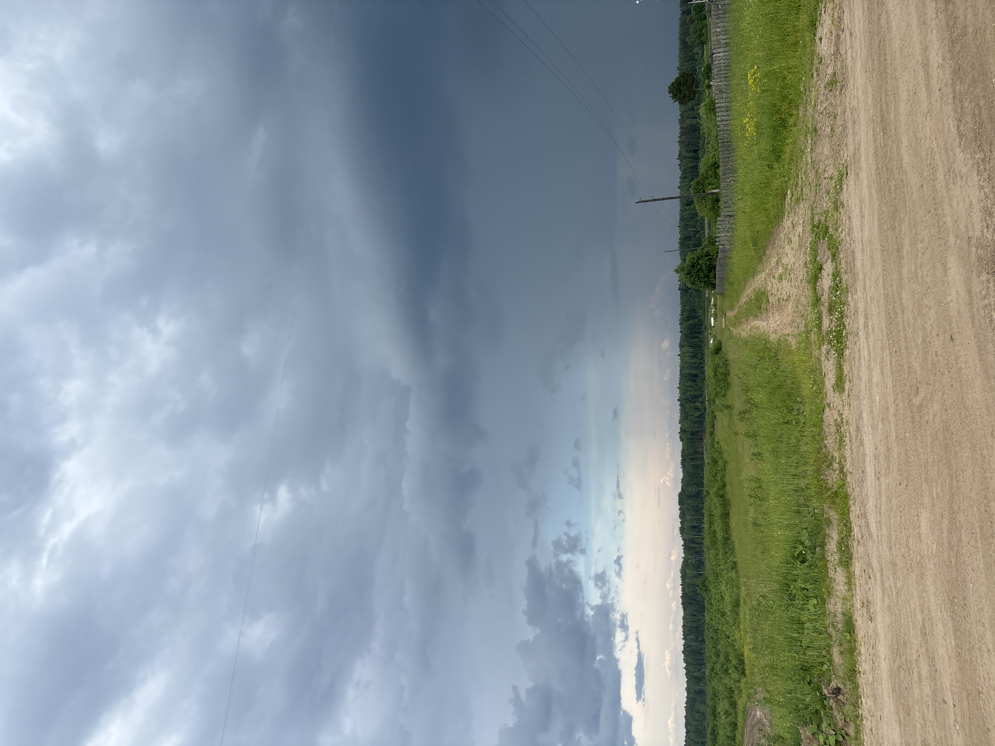 The image depicts a rural landscape under a stormy sky. In the foreground, a dirt road curves through a grassy field. In the middle ground, a line of trees forms a dense forest. In the background, the sky is dominated by dark, ominous clouds, suggesting an approaching storm. Power lines and poles are visible against the sky, indicating the presence of human infrastructure. The overall impression is one of an impending weather event in a natural setting. The location is identified as the city of Kurgan, Russia. There are no people or animals visible in the scene. The environment appears to be a field and forest, with a dirt road and utility poles.