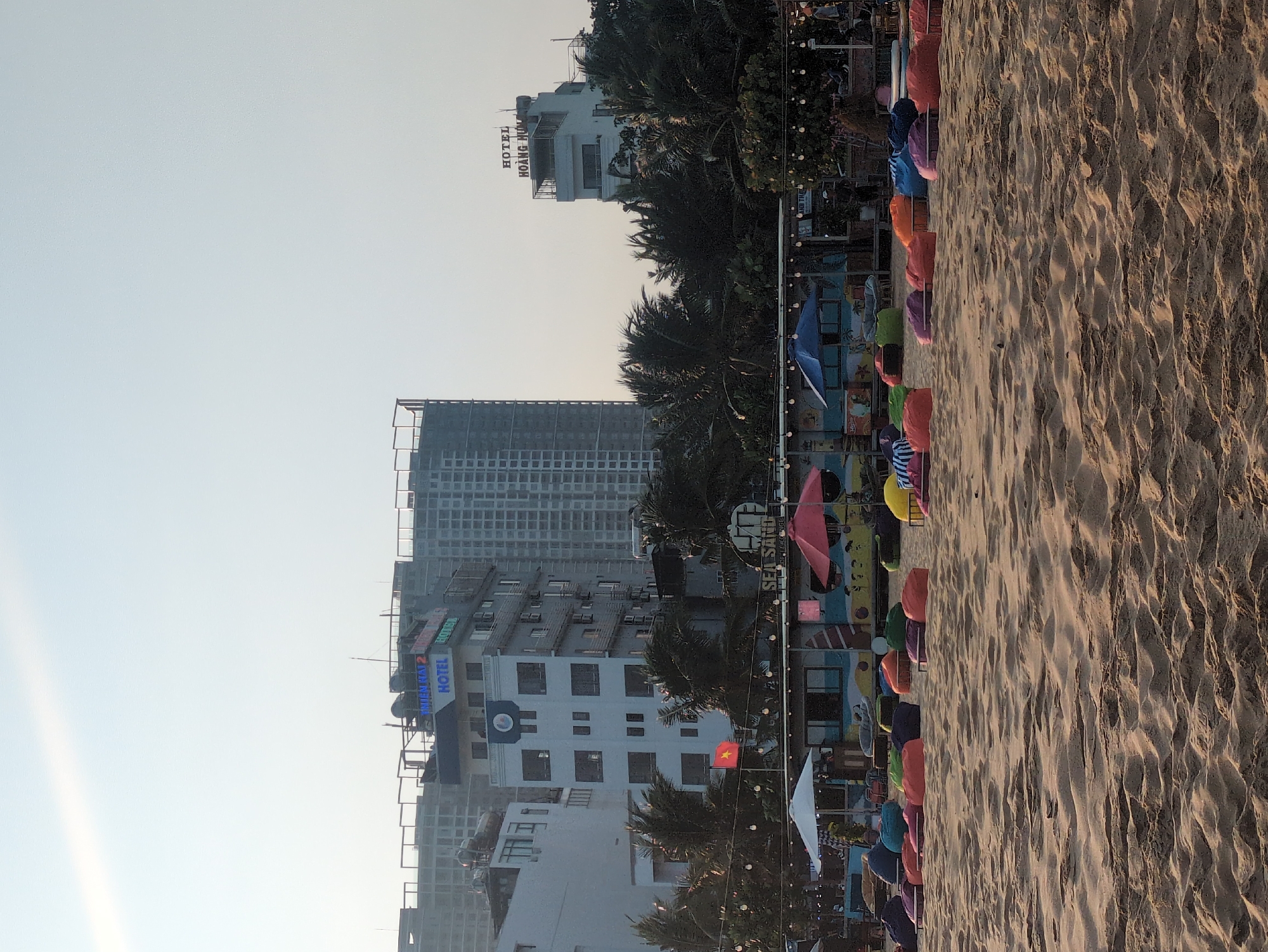 The image shows a beach scene in Quy Nhon Ward, Vietnam. The foreground is dominated by a wide expanse of sand. A line of colorful beach umbrellas and beanbag chairs is arranged along the edge of the beach. In the background, several buildings are visible, including hotels. One building is labeled "SEA SANDS." Another building is labeled "HOANG NHU HOTEL." A Vietnamese flag is flying in the middle ground. The sky is clear and light.