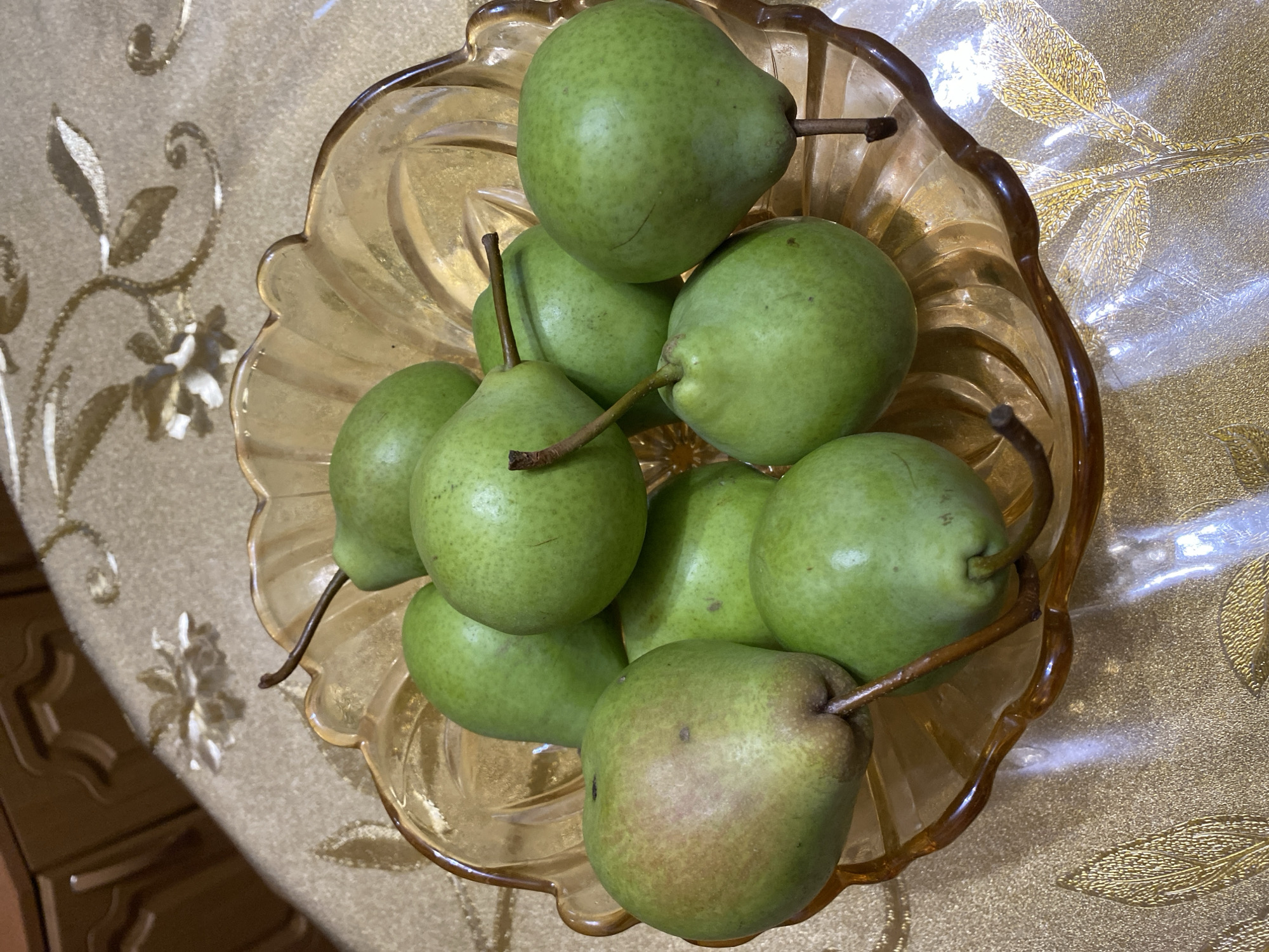 A bowl of green pears sits on a gold tablecloth with a floral pattern. The tablecloth is on a wooden table. The bowl is made of amber glass and has a scalloped edge. The pears are all green and have brown stems. Some of the pears have slight blemishes. The lighting in the image is dim and warm. The image appears to be taken indoors. The location is not discernible from the image.