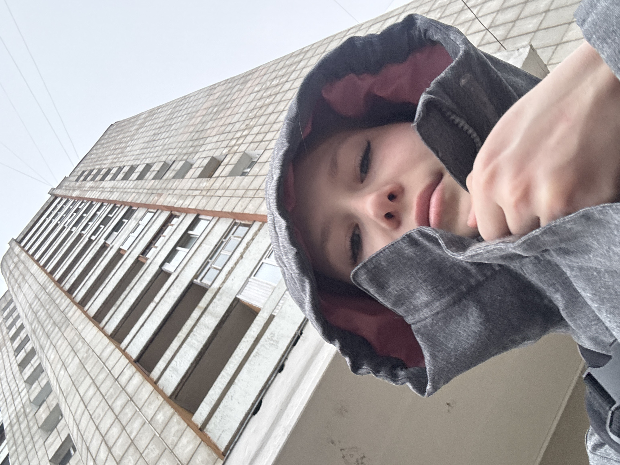 A young person wearing a gray hooded sweatshirt with a pink lining is the primary subject of the photograph. The camera is positioned looking upwards at the person, creating a low-angle shot. The background features a tall building with multiple floors and rows of windows. The building appears to be constructed with rectangular blocks, possibly concrete or tile, and has a somewhat aged appearance. The presence of utility lines suggests an urban environment. Based on the building style, this is likely a residential or commercial area. The style of the building is indicative of Soviet-era construction, which matches the city of Chaikovsky, Russia.