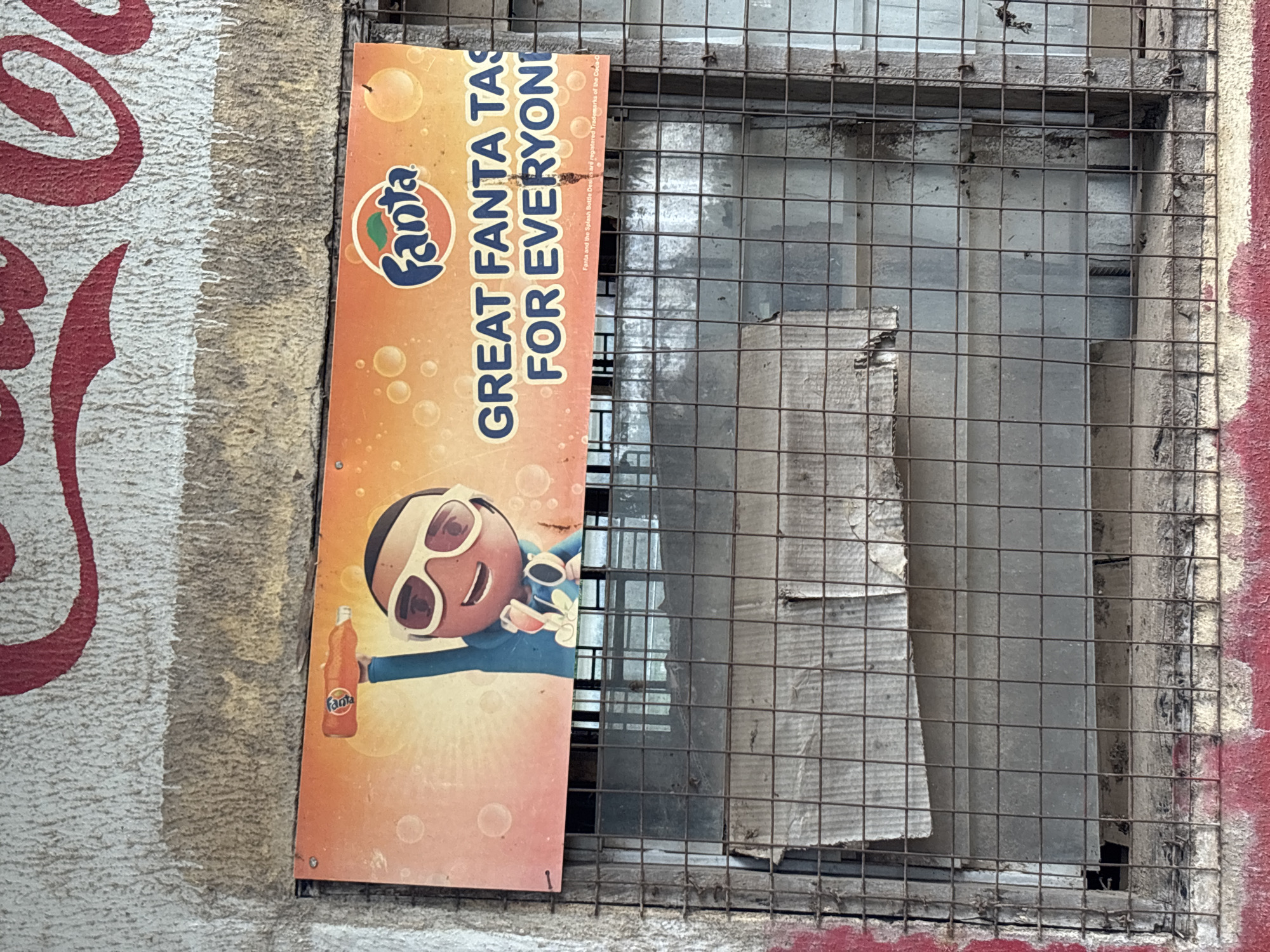 A Fanta advertisement is displayed on a corrugated metal sign attached to a textured, weathered wall. The sign features a cartoon character with large sunglasses holding a bottle of Fanta. Text on the sign reads "GREAT FANTA TASTE FOR EVERYONE." The sign is positioned above a window covered with a wire grate, behind which obscured structures are visible. The lower part of the wall is painted red and shows signs of wear and tear. The overall environment suggests an outdoor setting, possibly in a commercial or urban area, given the advertisement and the building's condition. The image was taken in Jos, Nigeria.
