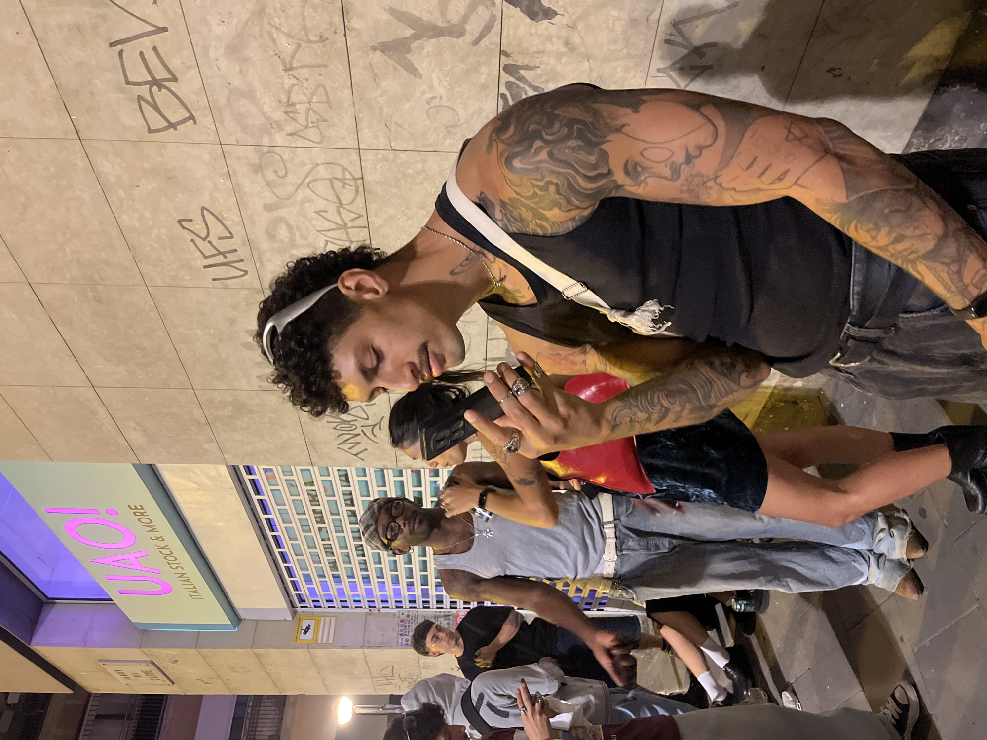A group of people stand on a sidewalk in Barcelona, Spain. The location is identifiable by the "UAO! Italian Stock & More" store sign visible in the image. The group includes a heavily tattooed man in a black tank top using a cellphone held by a person wearing a red top and dark miniskirt. Another man in a grey tank top and checkered cap stands adjacent to them. Further back, there are additional people, one wearing a black t-shirt. The sidewalk is marked with graffiti. The lighting is ambient and suggests it is nighttime.