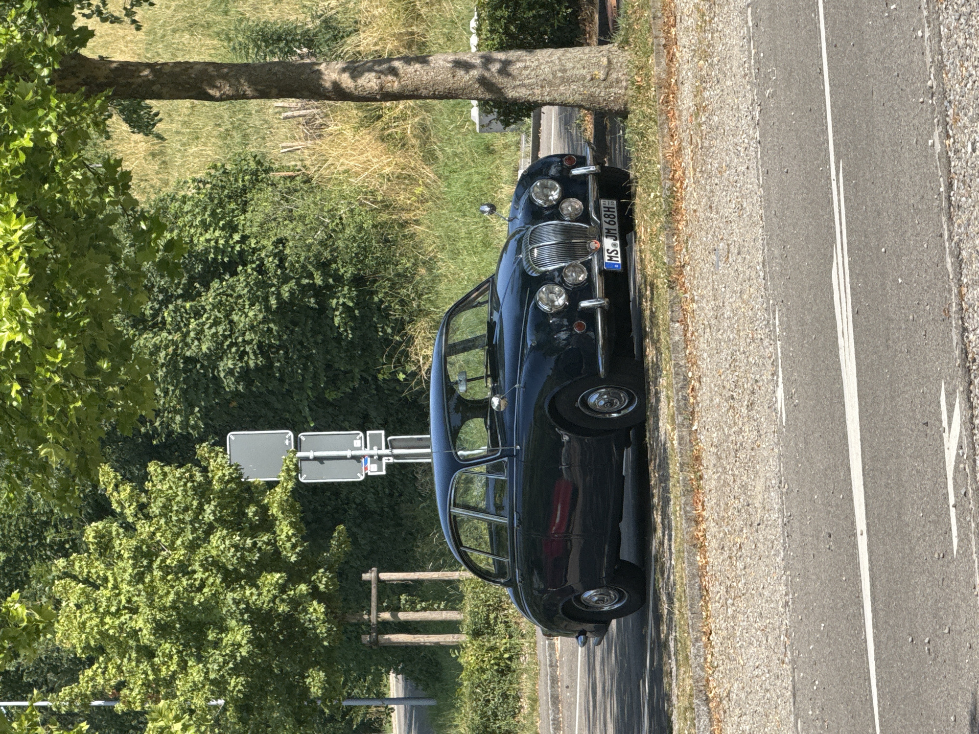 A dark-colored, vintage car is parked on the side of a paved road. The car has a chrome grille and headlights. The license plate is visible and reads "MS JM 68H". The car is positioned with its front facing to the left of the frame, and the perspective is from slightly above and to the right of the car.
The scene is outdoors, with a grassy area and trees to the left of the car. Sunlight is casting shadows, suggesting it is daytime, likely afternoon given the angle of the light. The weather appears to be clear and sunny. In the background, there are more trees and vegetation. A tree trunk is prominently visible in the left foreground. Traffic signs, one with a rectangular grey face, are also visible behind the car. The road has white lines marking the lanes. The ground next to the road is covered in gravel and dry leaves. The overall setting appears to be a roadside in a rural or semi-rural area. There are no people visible in the image. The image conveys a sense of a quiet, sunny day.