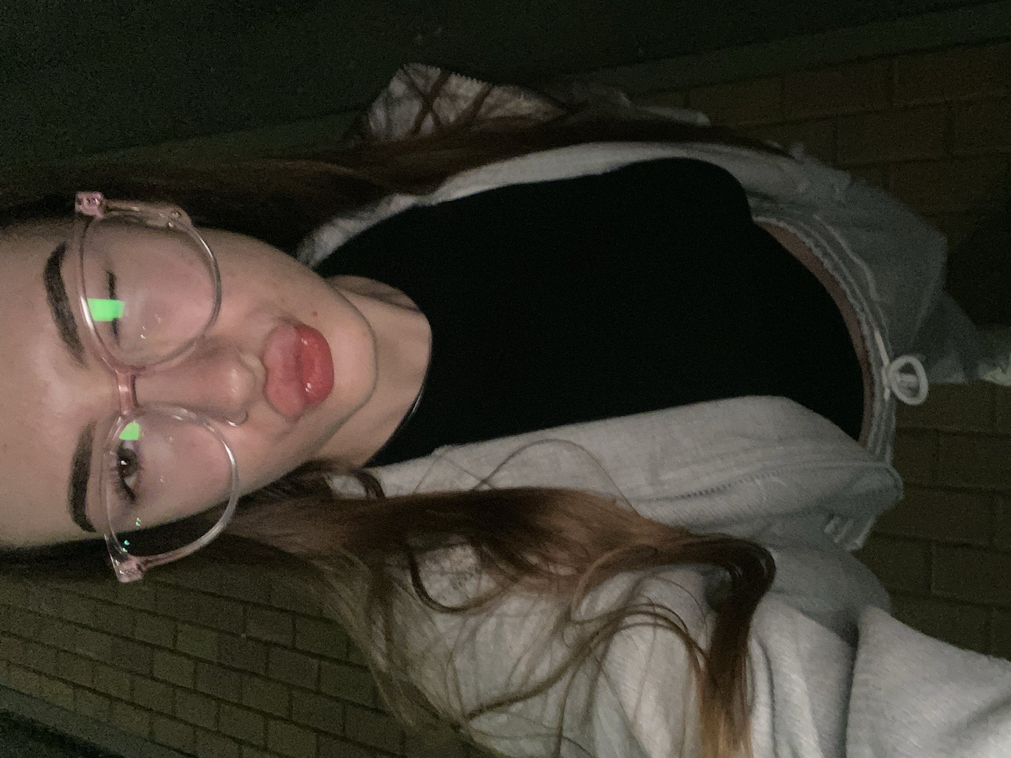 A young female, with long brown hair, is captured in a close-up, slightly elevated selfie. She has light skin, full eyebrows, and is wearing clear, pink-framed cat-eye eyeglasses with a visible green light reflection in the right lens. Her lips are full, puckered into a pout with a reddish-pink gloss, and she has a small metallic ring piercing on her left nostril. She is wearing a black top, a light grey open hooded sweatshirt, and light grey sweatpants with a white drawstring visible. The background is dark, indicative of nighttime or low-light conditions. A dark paved surface, possibly brickwork or tiles, is visible to the left and bottom of the frame. The location is in the city of Unknown, Unknown.