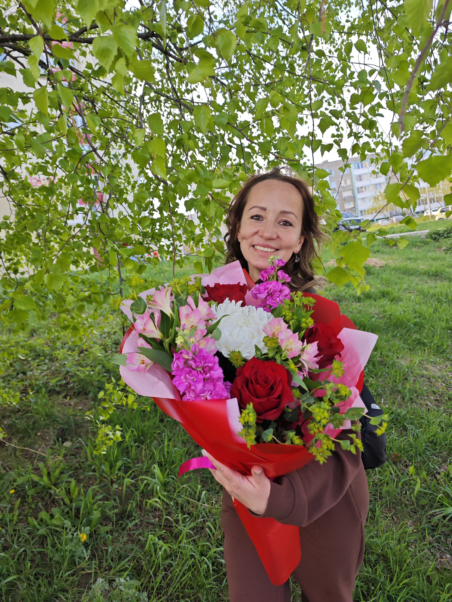 The image shows a woman holding a large bouquet of flowers, smiling at the camera. She stands in a grassy area with a tree behind her. The bouquet includes red roses, pink alstroemeria, pink stock flowers, and a white chrysanthemum. The bouquet is wrapped in pink and red paper and tied with a pink ribbon. The woman has brown hair and is wearing a brown sweatshirt. The background includes a building and some parked cars, suggesting an urban setting. The location is Chaikovsky, Russia.